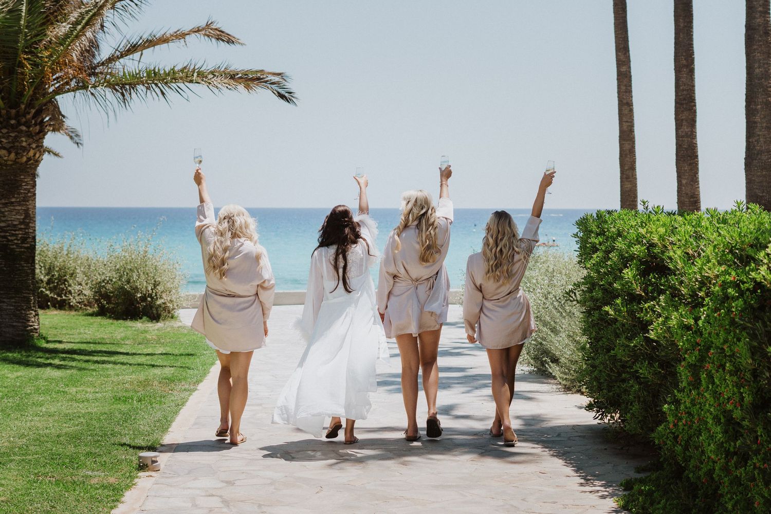 Bridal party in light dresses celebrates on palm-lined oceanfront path.