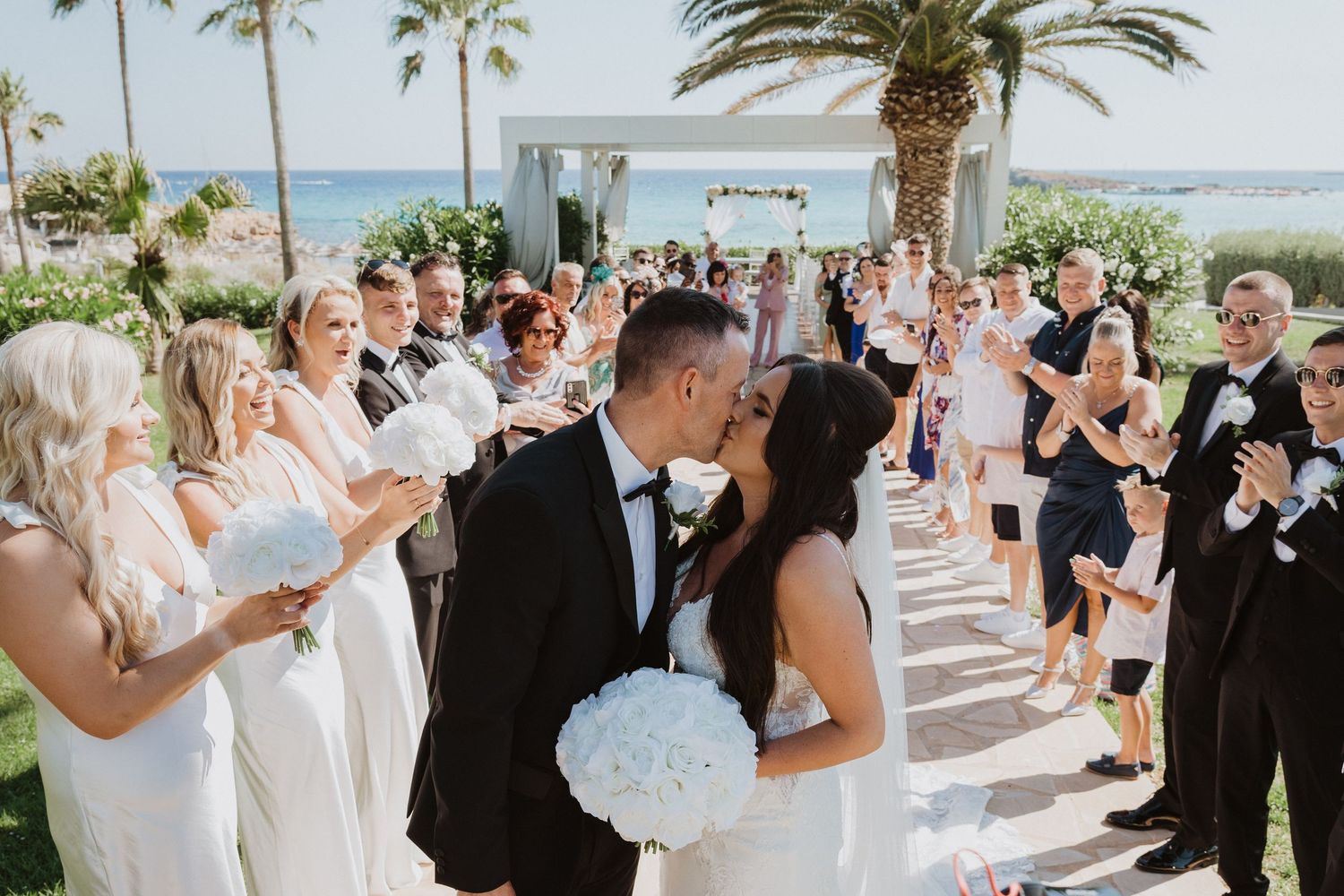 Elegant outdoor wedding ceremony with guests gathered along a palm-lined walkway.