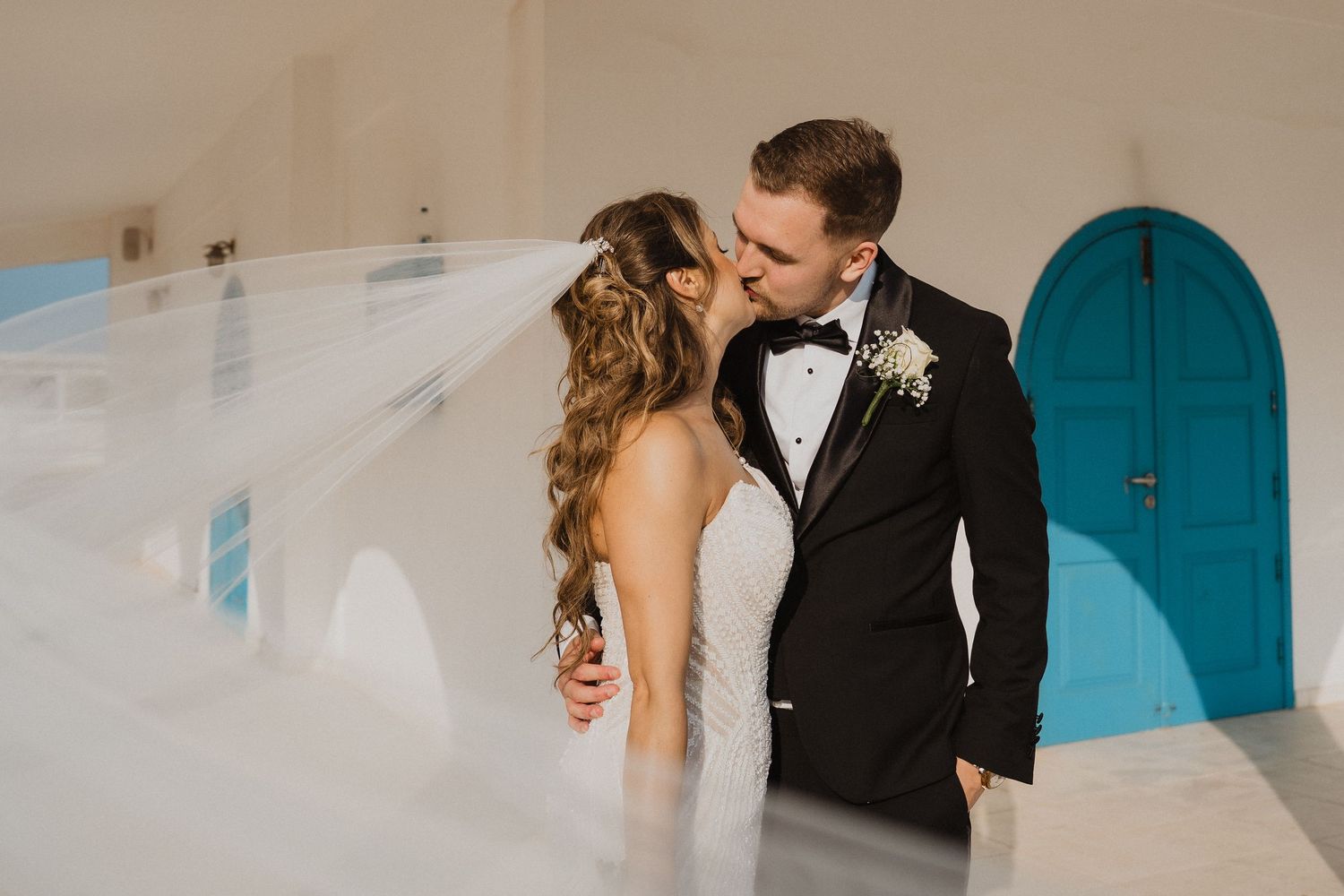 Bride and groom kiss near white wall with turquoise door as brides veil billows in the breeze.