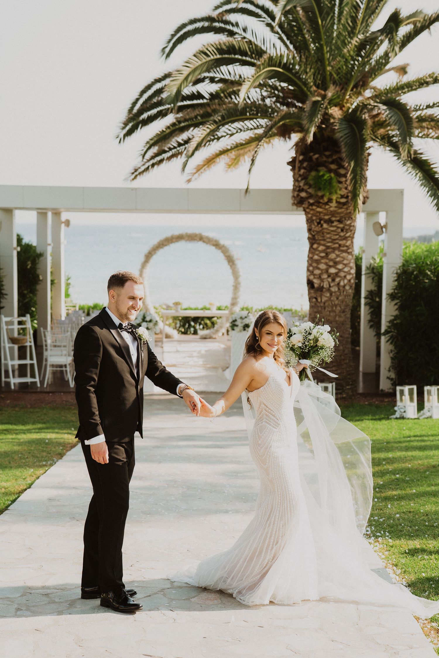 Bride in beaded gown holds grooms hand with flowing veil near palm tree at seaside venue.
