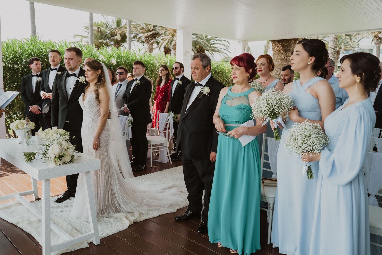 Wedding party stands at altar during outdoor ceremony with bridesmaids in blue and teal dresses.