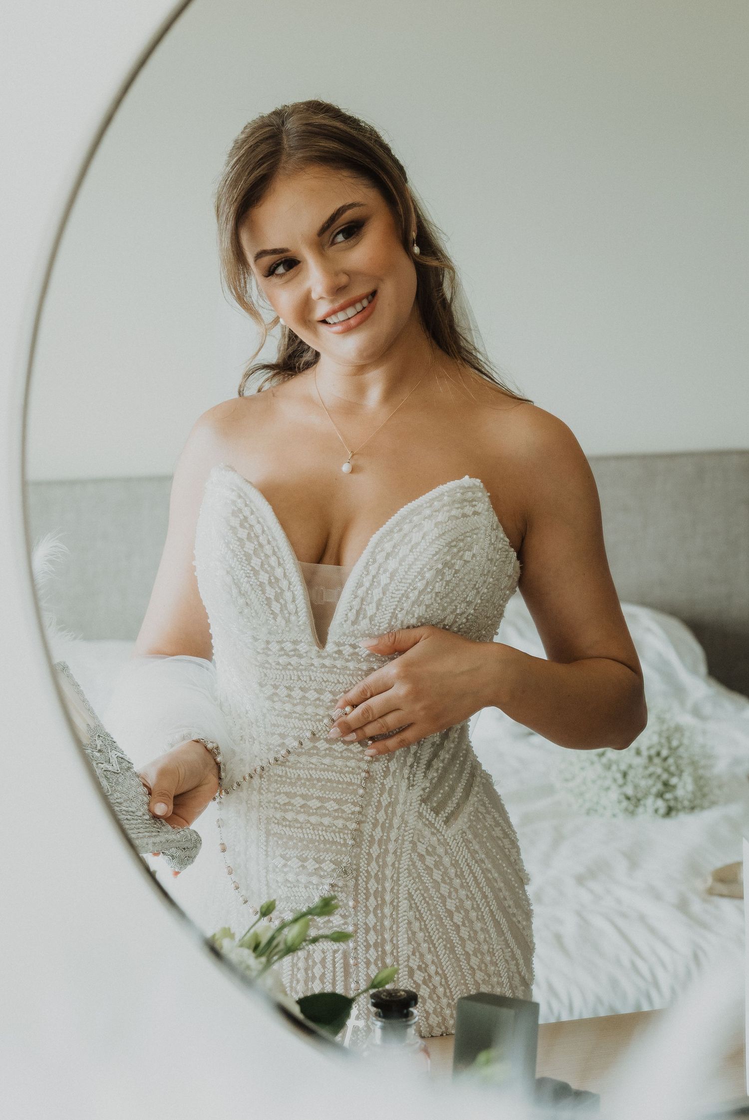 Beautiful bride in fitted beaded strapless wedding dress smiles at her reflection in a round mirror.