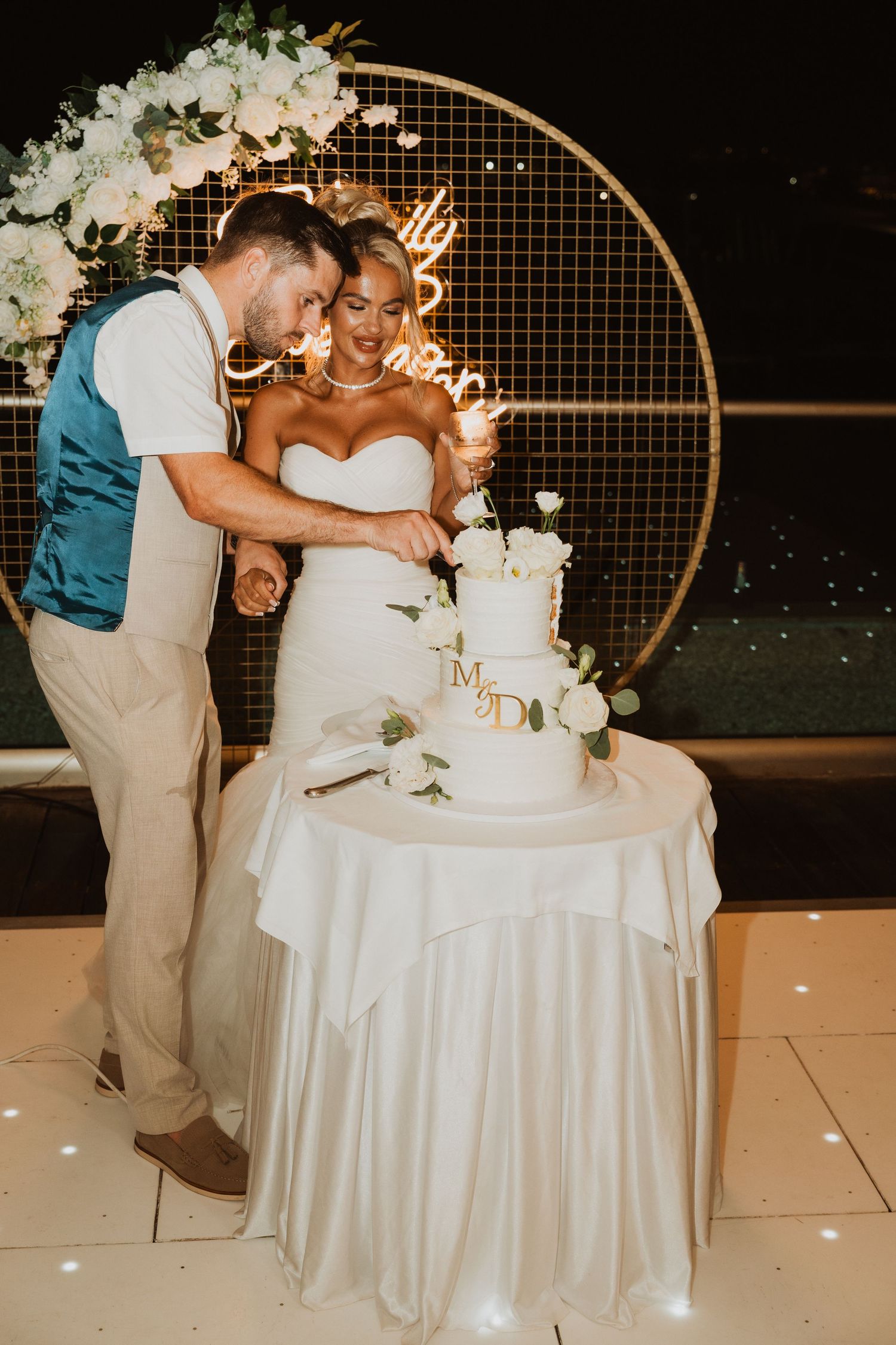 Bride and groom cutting white three-tier wedding cake decorated with roses at elegant outdoor night reception.