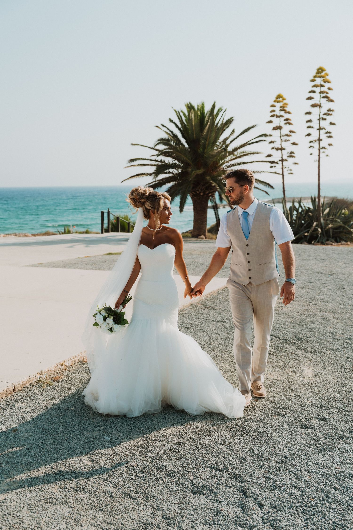 Bride and groom holding hands walking by the ocean with palm trees, bride in white mermaid gown, groom in tan suit.