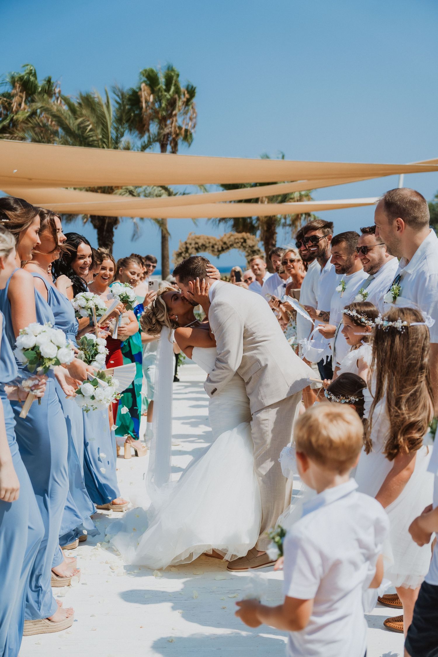 Bride and groom share a romantic kiss at outdoor beach wedding ceremony with guests in blue dresses and white attire.