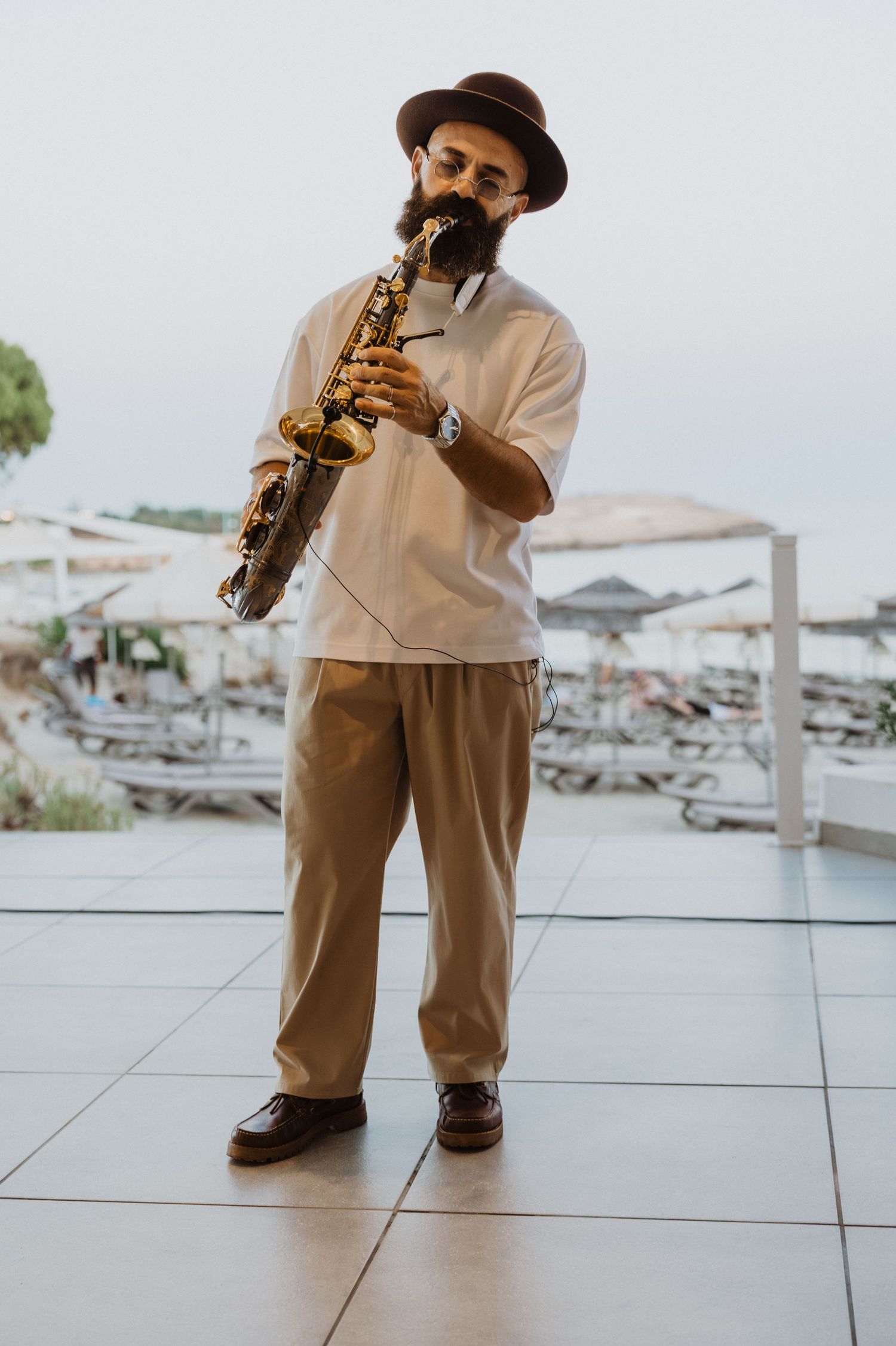 Musician in a hat and light-colored suit playing saxophone on an outdoor terrace.