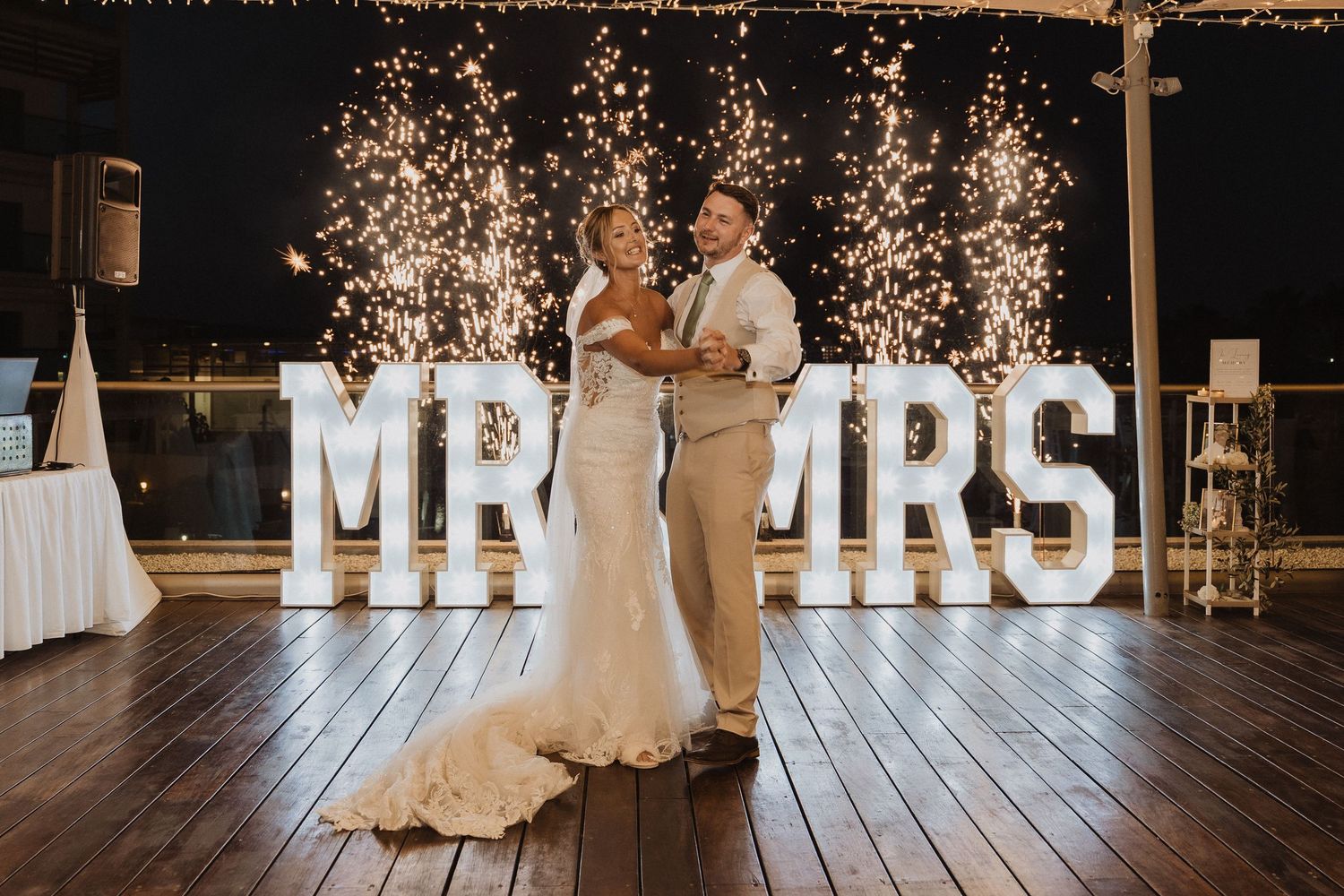 A couple shares their first dance at a wedding reception with illuminated MR & MRS letters glowing in the background.