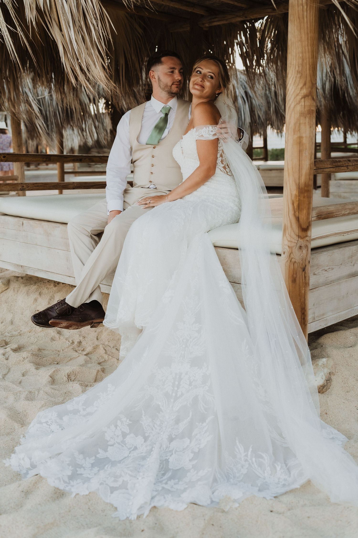 A romantic beach wedding couple poses under a thatched palapa with a stunning lace wedding dress trailing in the sand.