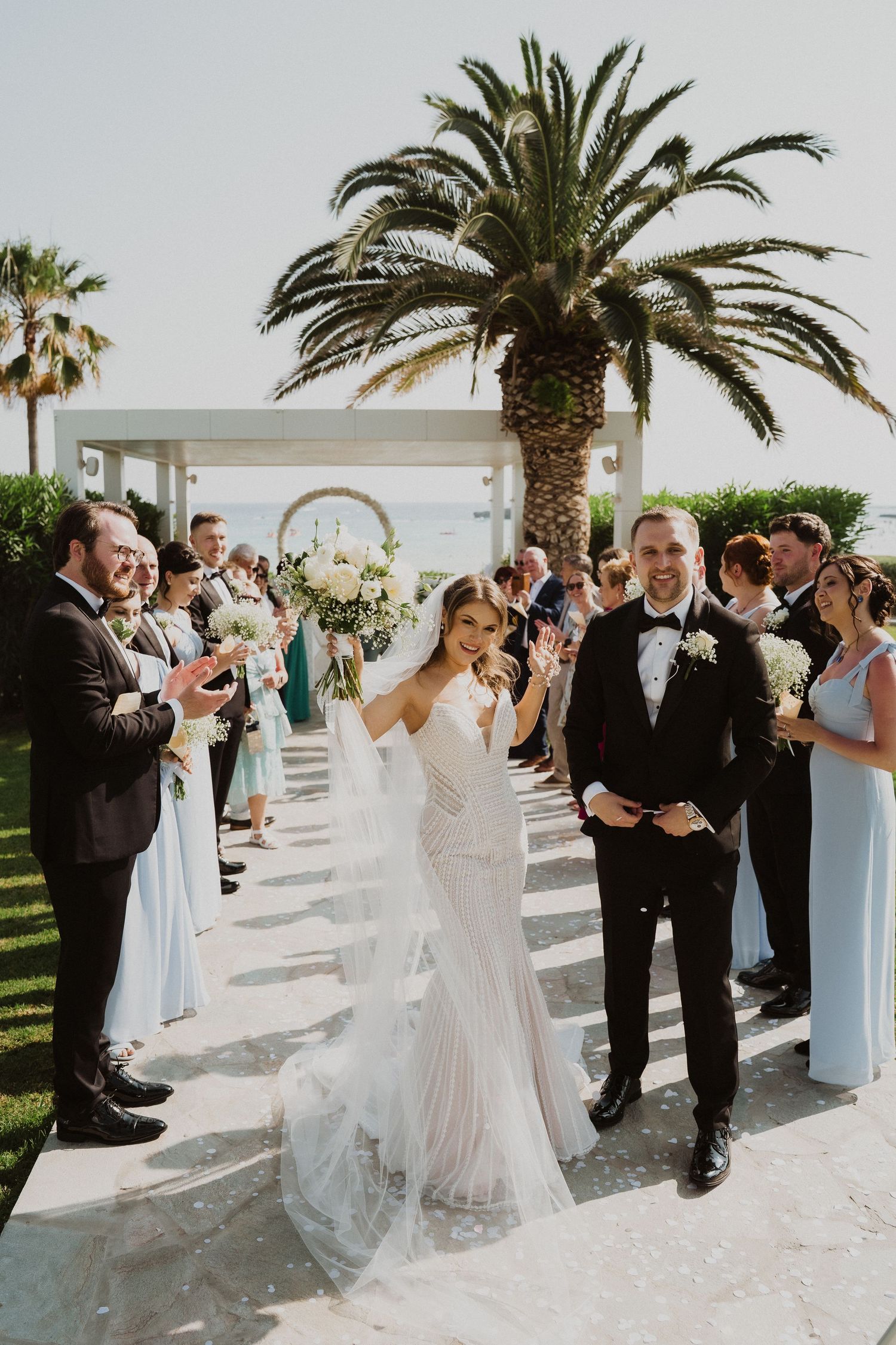 Newlyweds walk down aisle surrounded by guests in light blue dresses and black tuxedos.