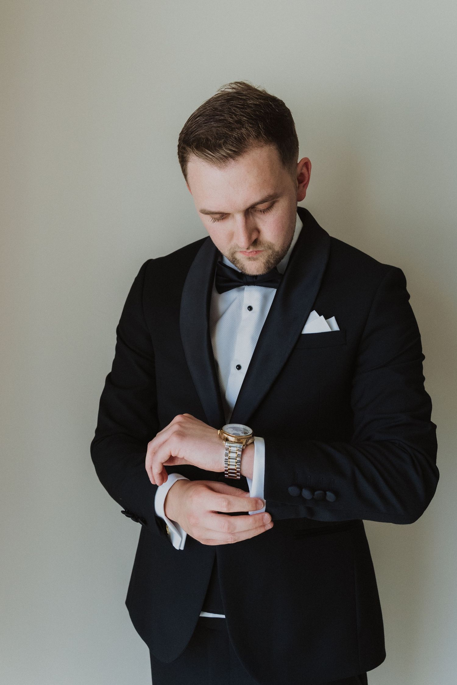 Elegant groom in black tuxedo adjusting gold watch cufflinks before wedding ceremony.