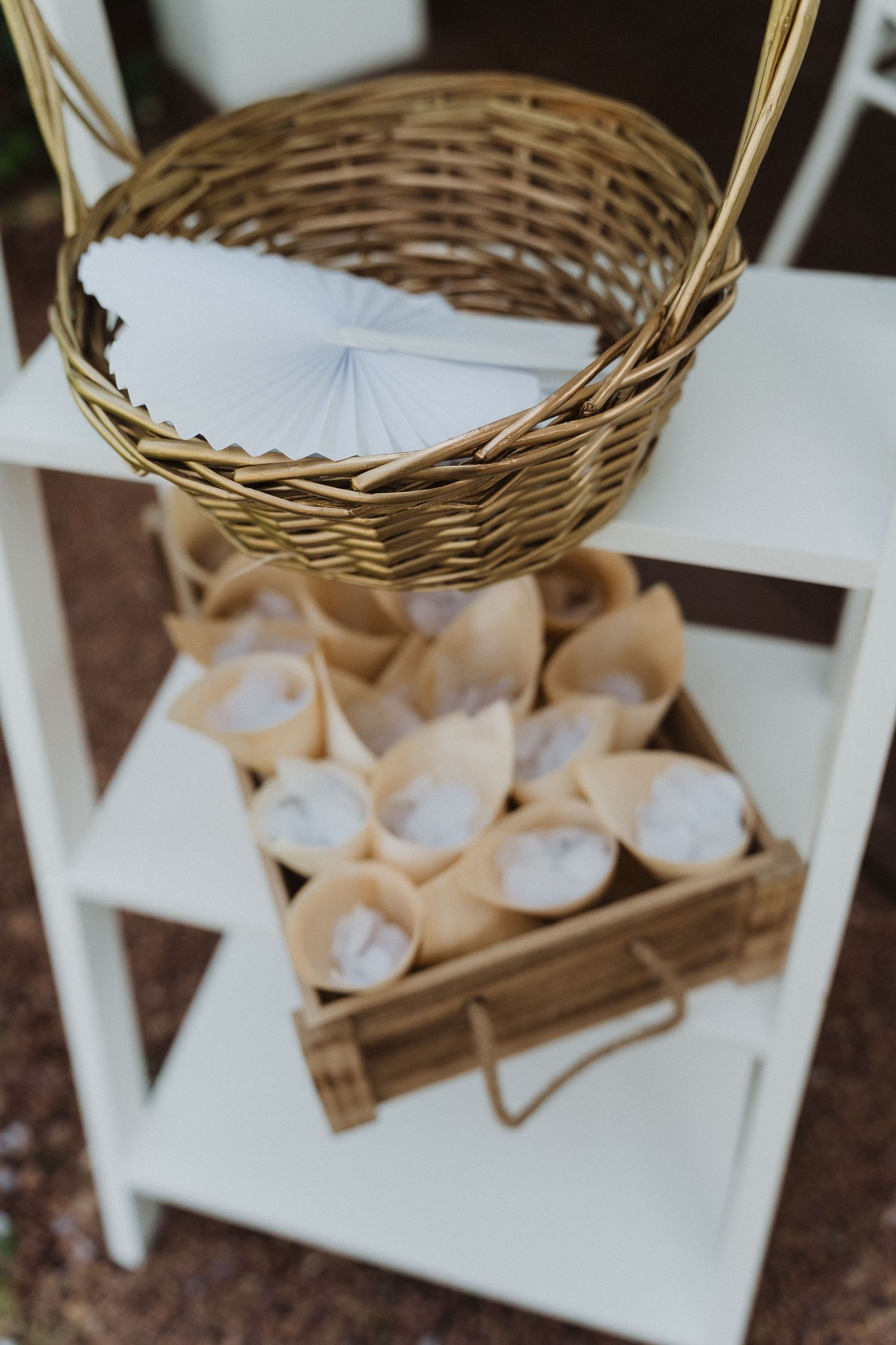 Rustic wedding ceremony decor featuring wicker basket and kraft paper cones on a white shelf.