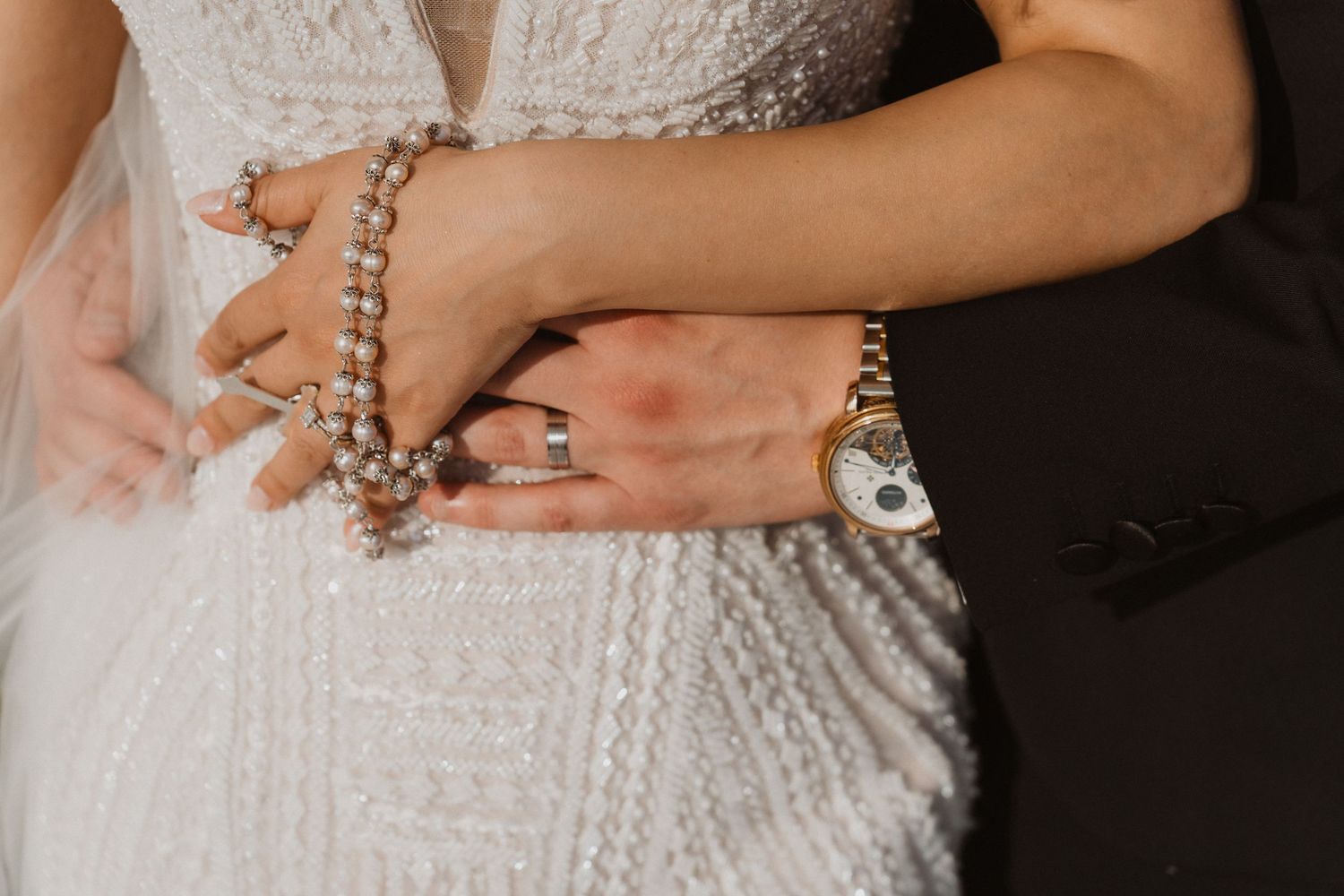 Close-up of bride and groom's hands, bride wearing pearl bracelet and diamond ring, groom wearing gold watch.