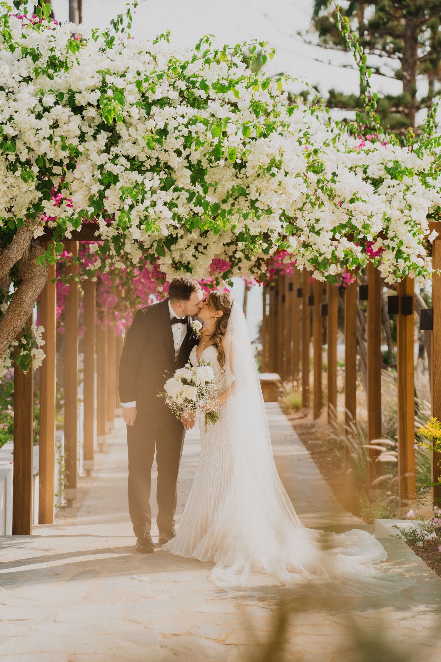 Bride and groom kissing under a floral pergola with white and pink bougainvillea blooms at an outdoor wedding.