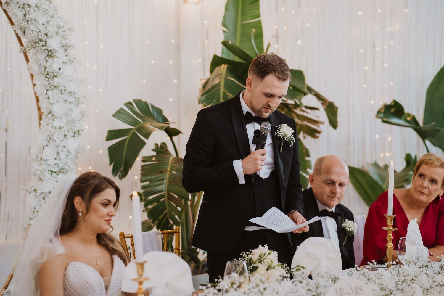 Groom in black tuxedo delivering wedding speech at head table decorated with white flowers and gold candles.