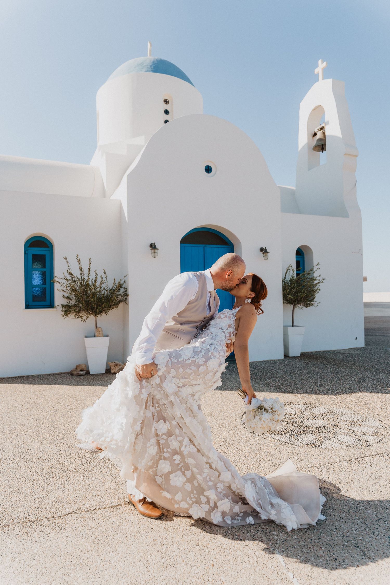 Traditional white Greek church provides backdrop for wedding photos with blue domed roof.