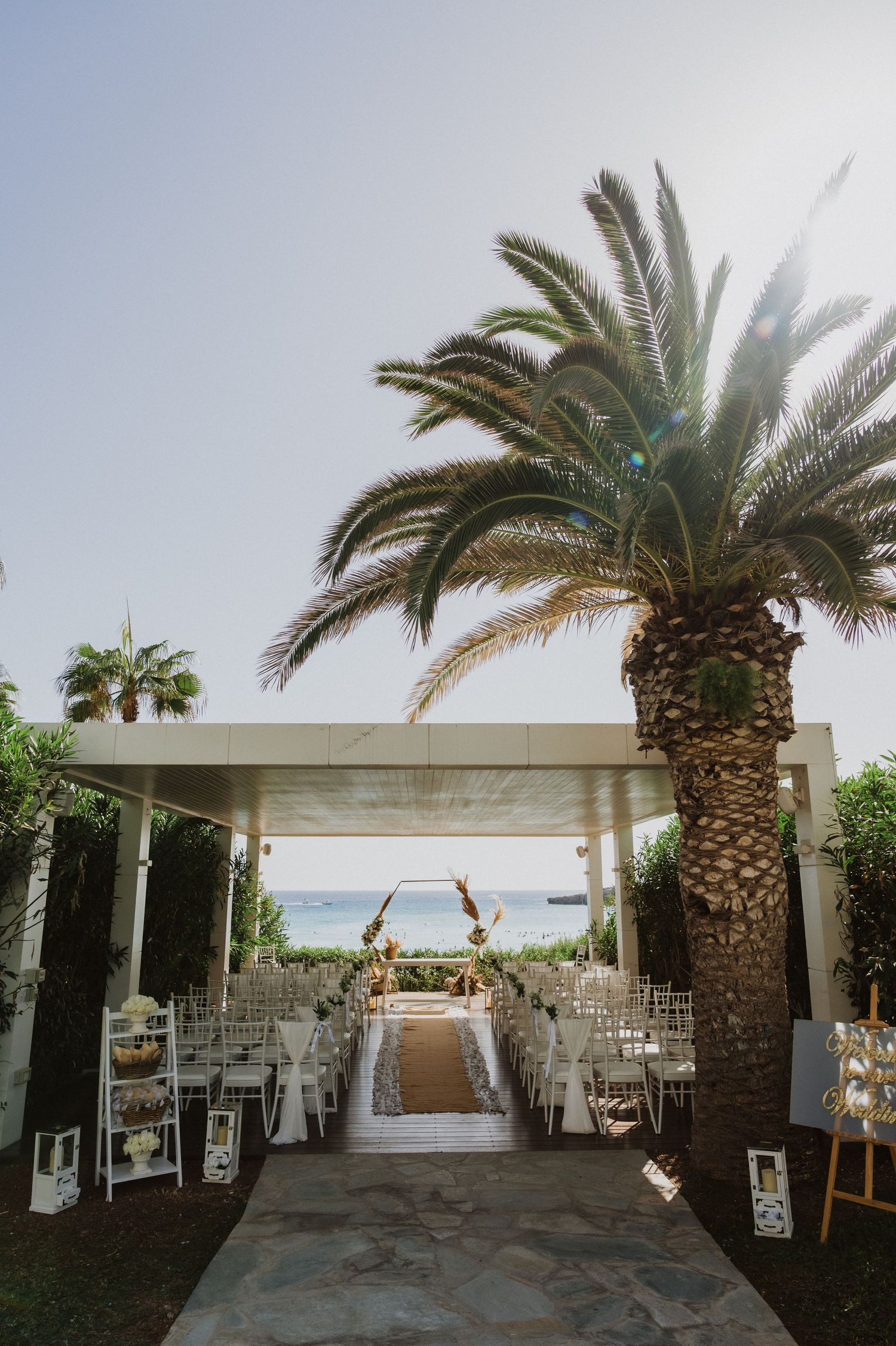 Wedding ceremony aisle with white chairs and palm trees leading to an ocean view arch.