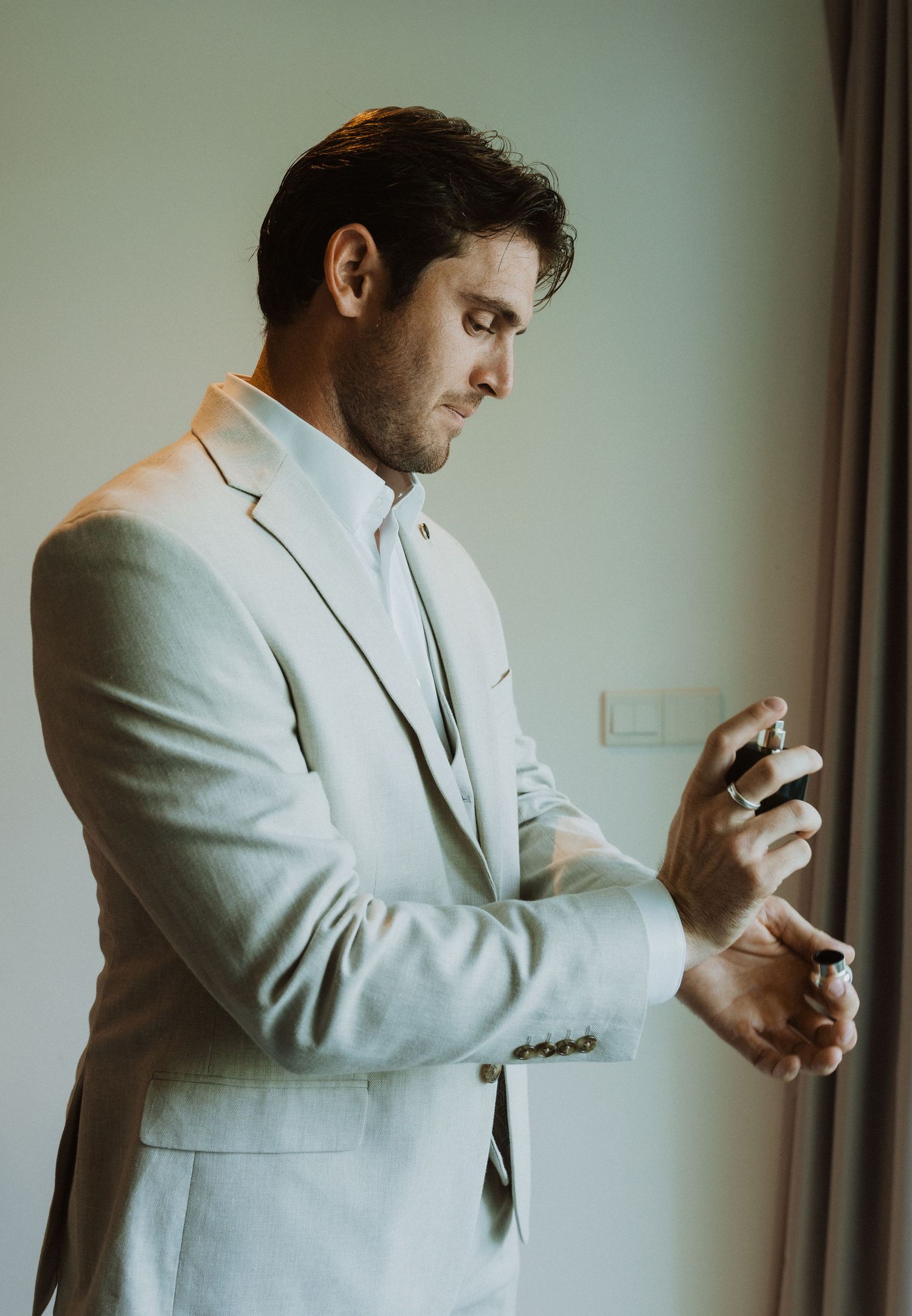 Person in light gray suit applying cologne near window.