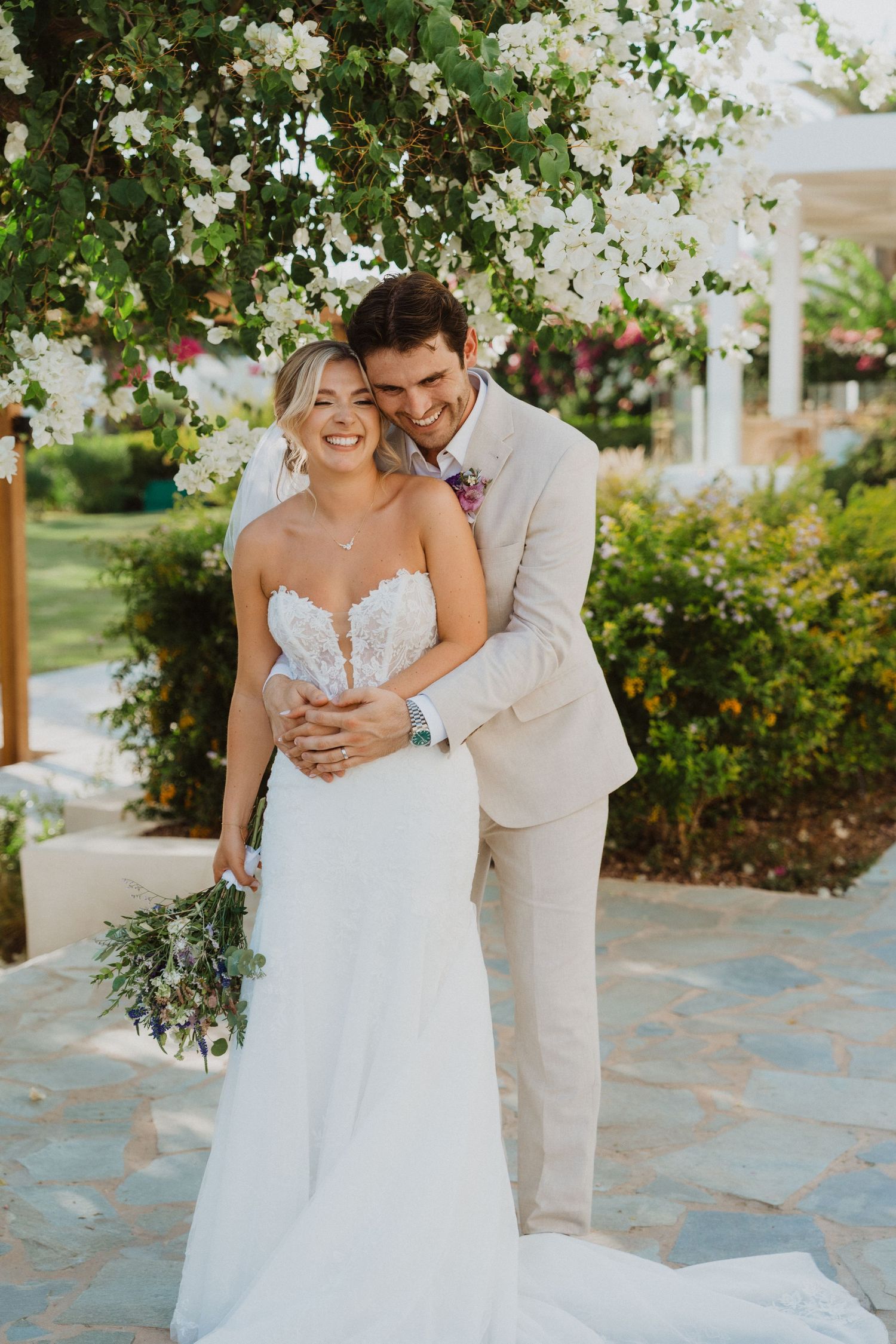 Wedding couple shares candid moments under blooming white flowers and greenery.