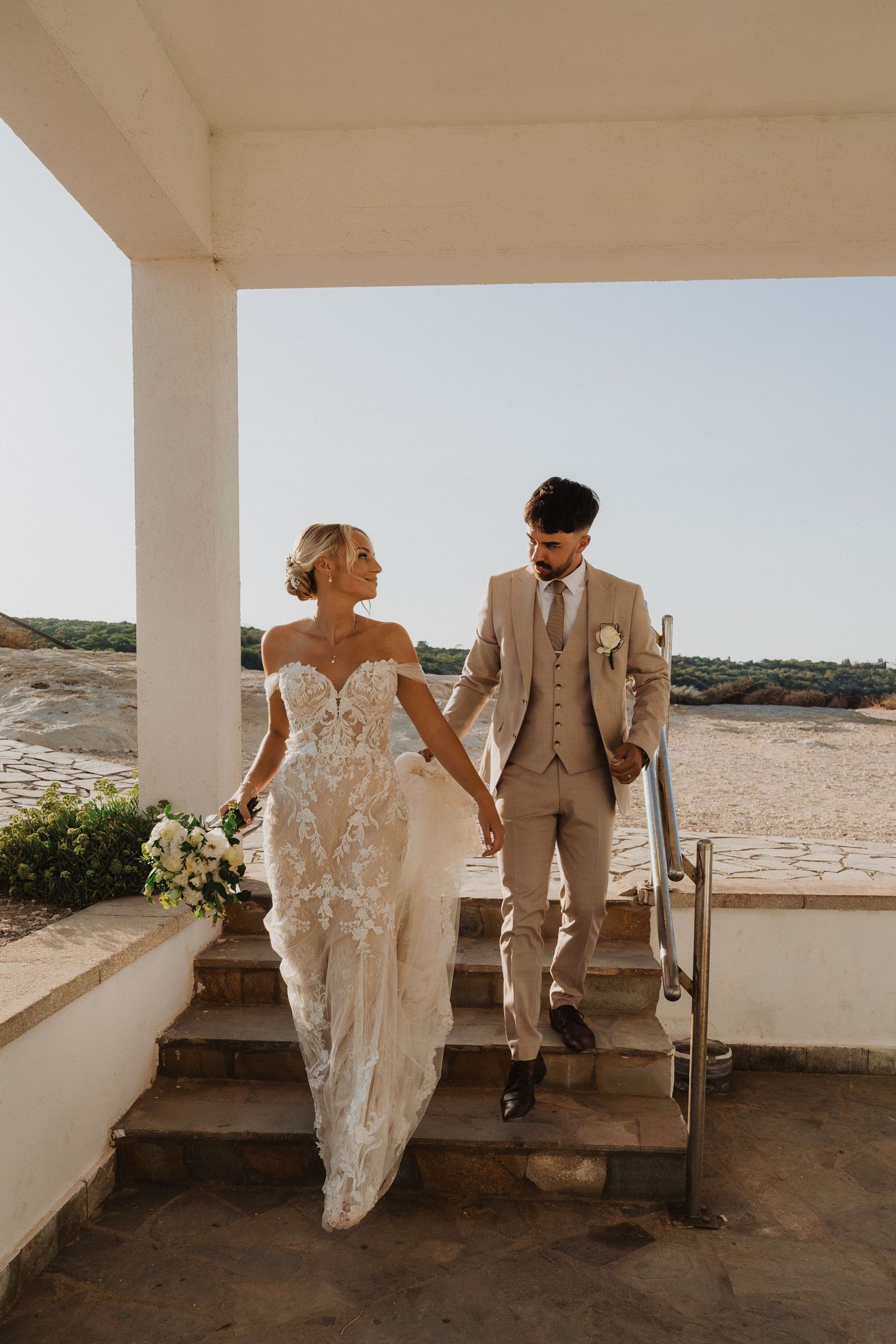 A romantic wedding couple poses against white columns with ocean views in their elegant attire.