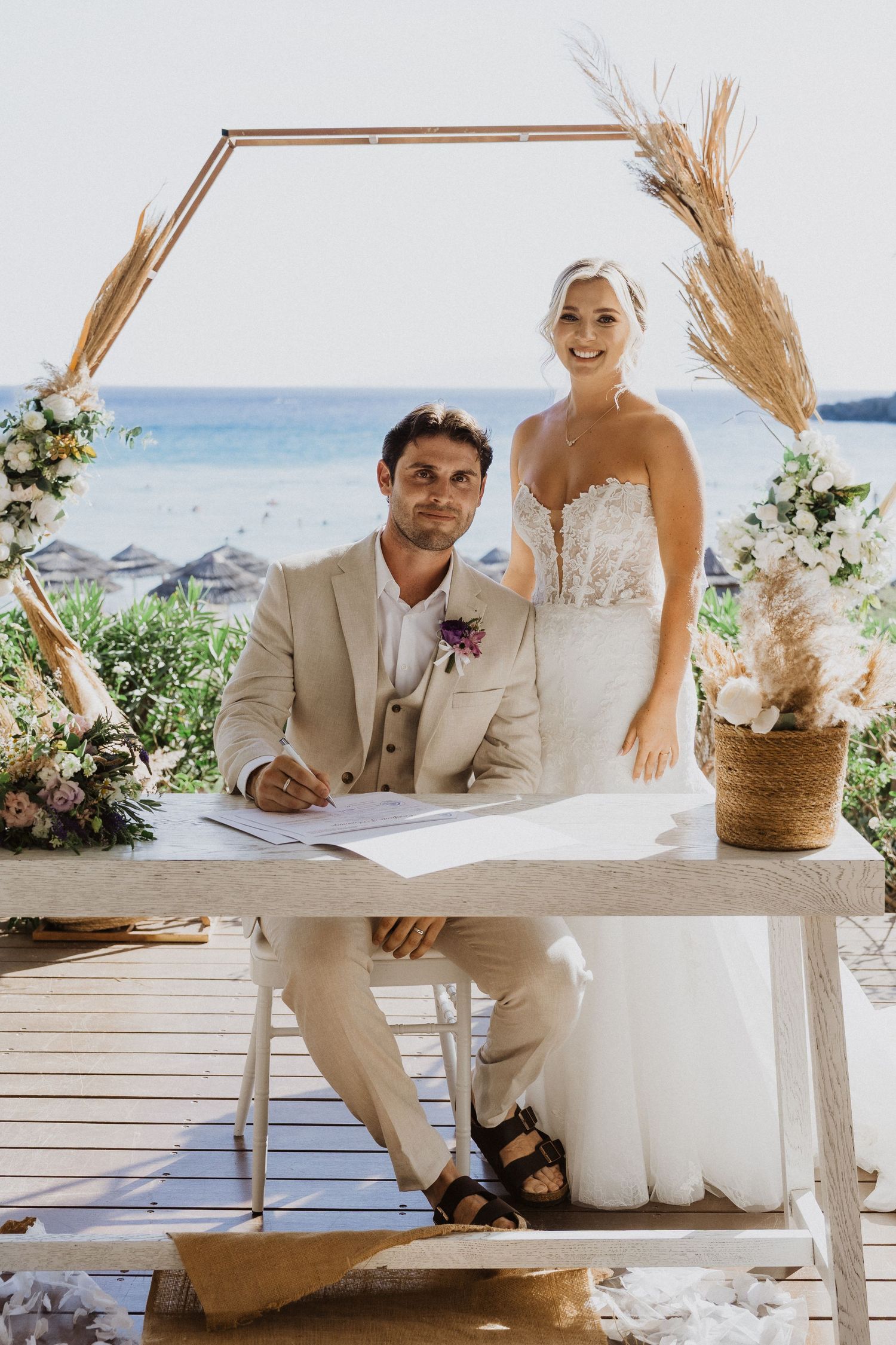 Beachfront wedding ceremony setup with rustic wheat decorations and white wooden furniture.