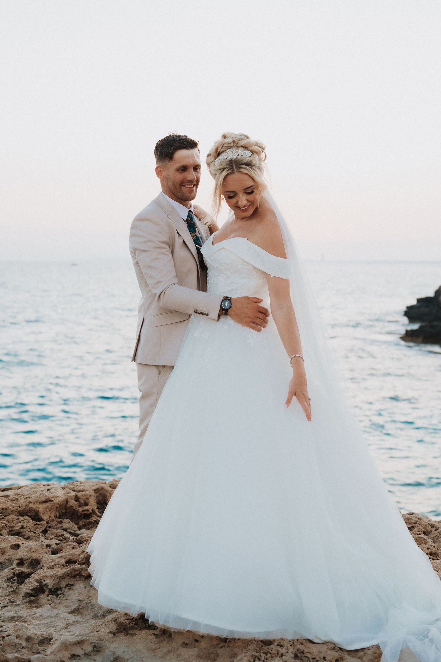 A bride in a white ballgown and groom in beige suit embrace on rocky cliffs overlooking the ocean at sunset.