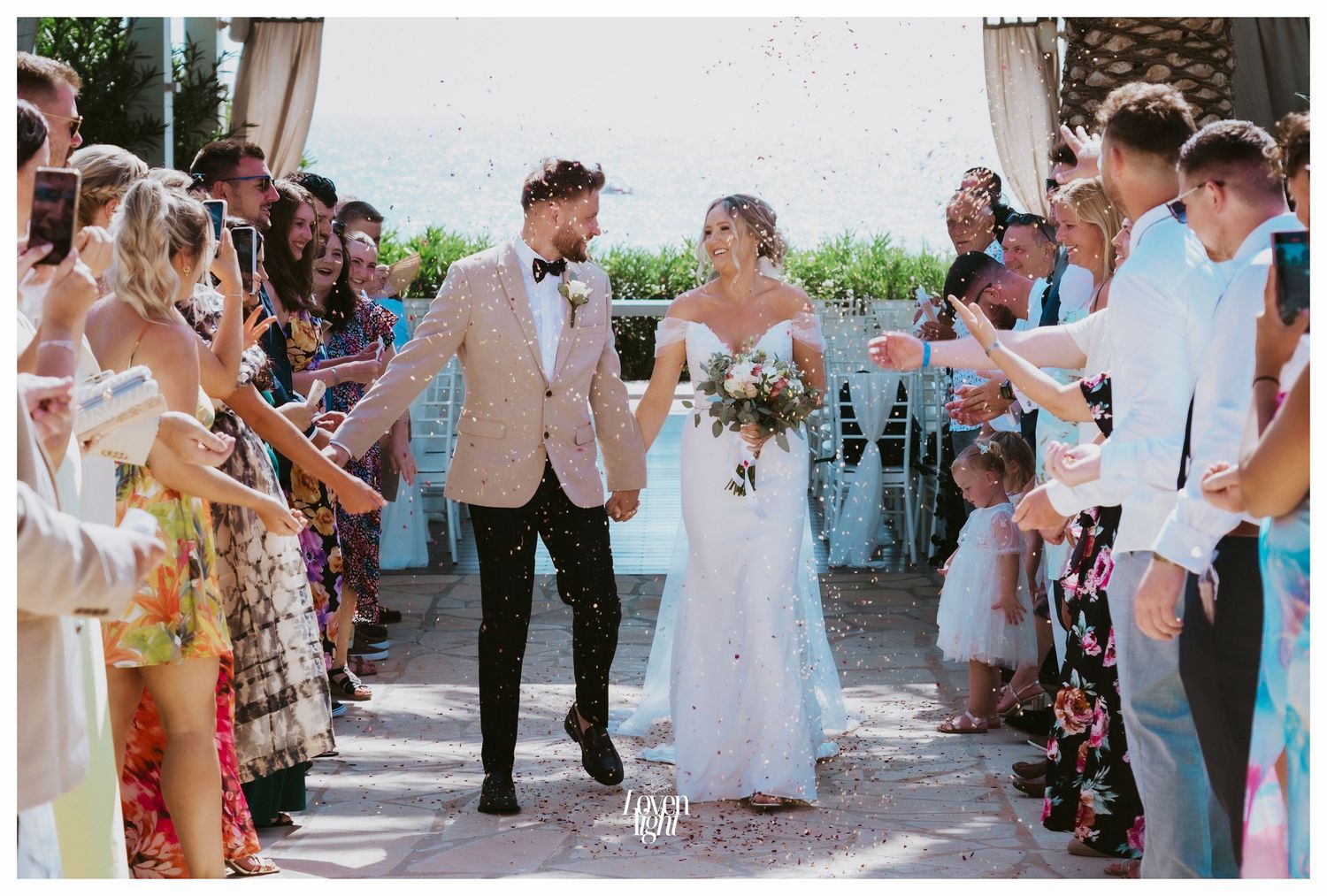 A couple walks down an outdoor wedding aisle while guests cheer and celebrate on both sides.