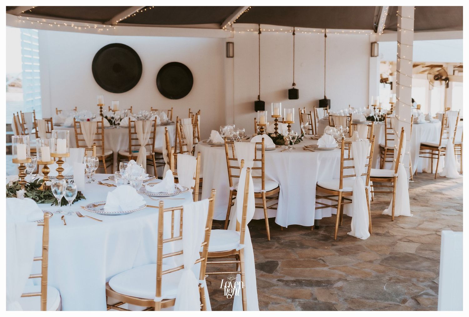 Reception venue with white-clothed tables and chiavari chairs arranged under a covered pavilion.