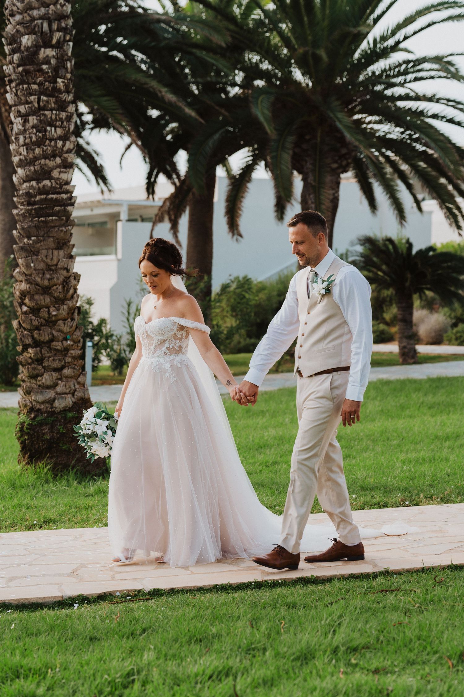 A couple in wedding attire walk hand in hand on a pathway surrounded by palm trees at a tropical venue.