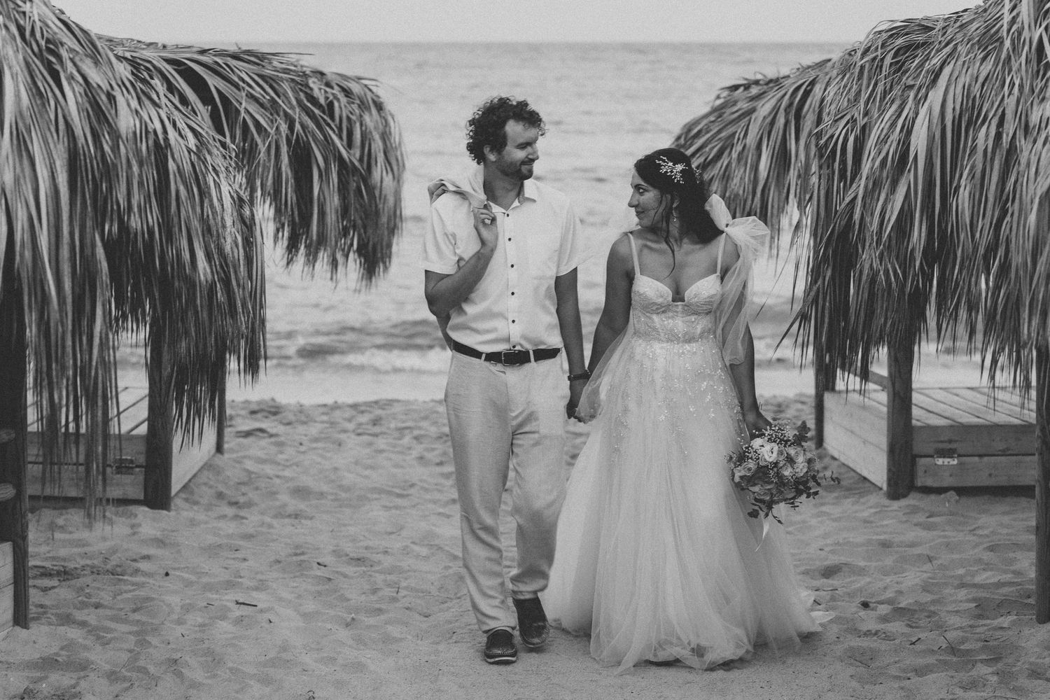 A black and white beach wedding photo of a couple walking between thatched palapas.