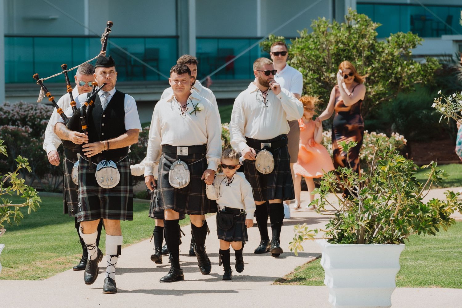 Traditional Scottish wedding procession with men and child in kilts walking down path.