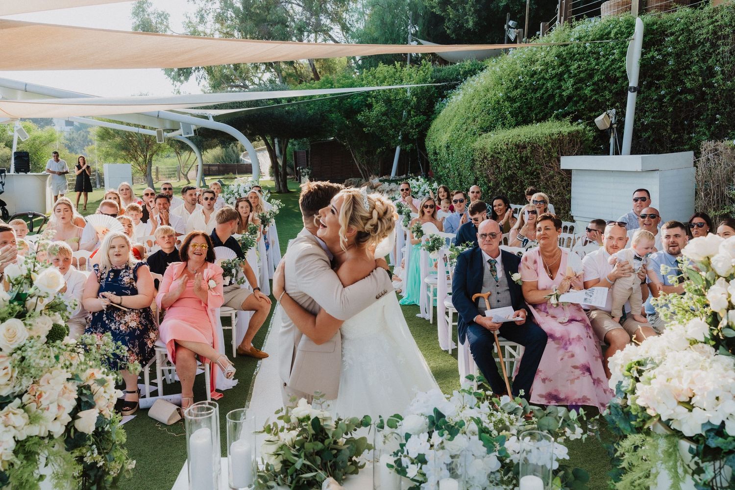 Outdoor garden wedding ceremony with lush floral arrangements and guests watching a couple embrace at the altar.