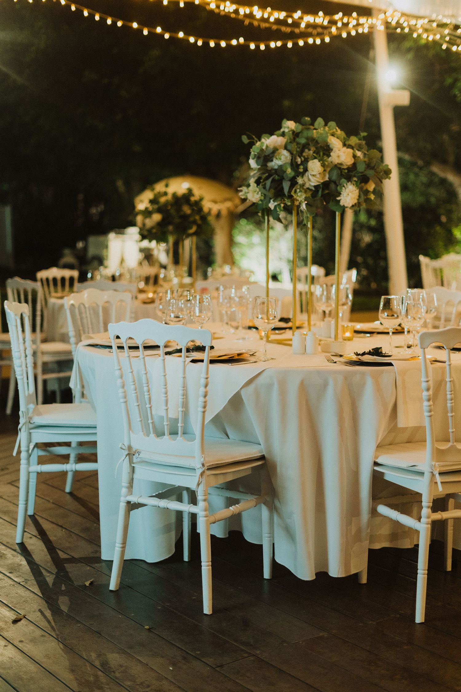An elegant outdoor wedding reception setup with white chiavari chairs and tables under romantic string lights.