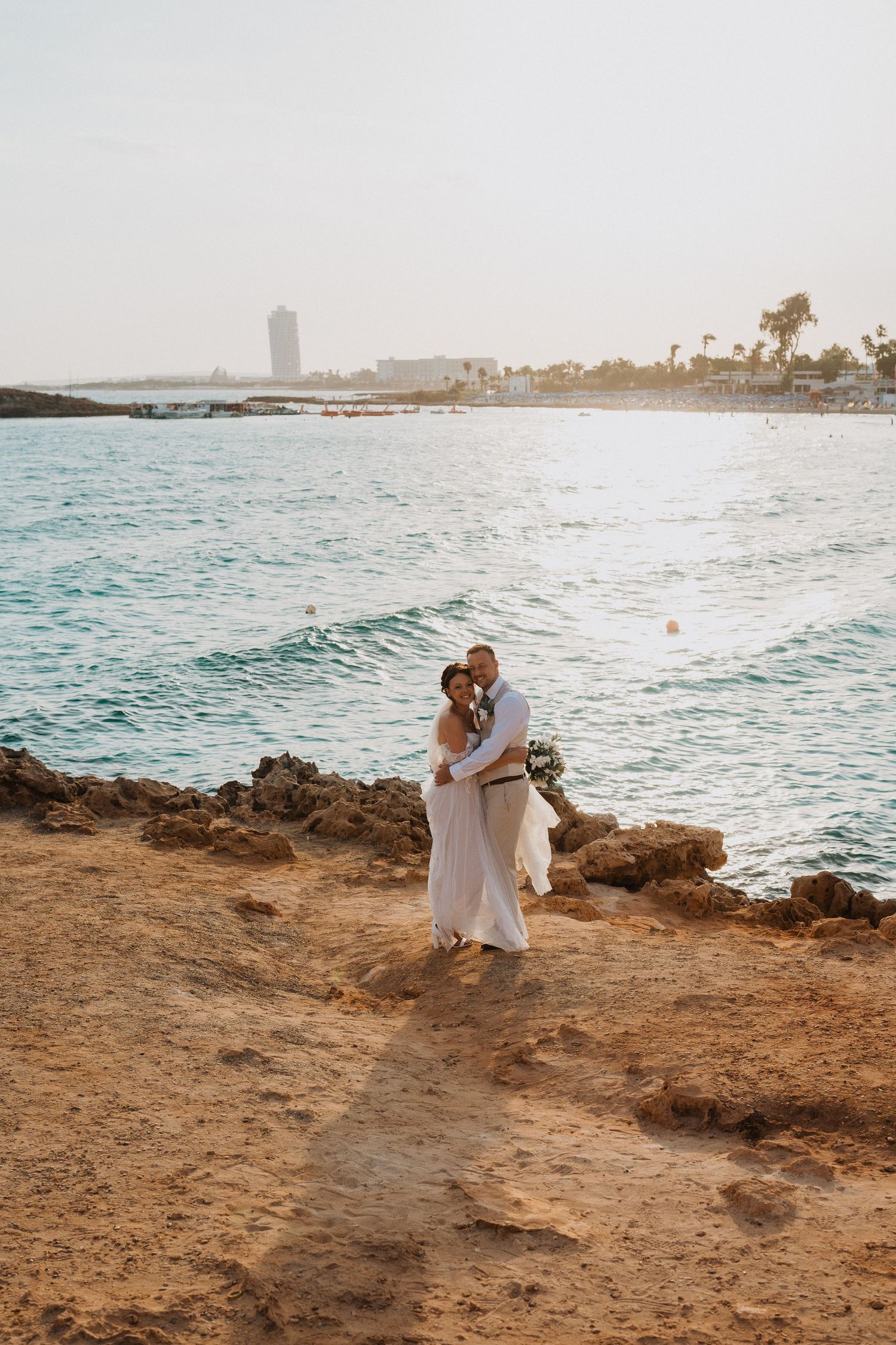 Couple in white embraces on rocky coastal cliff overlooking turquoise waters and distant cityscape at sunset.