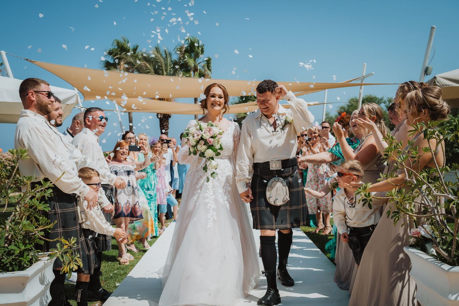 Beach wedding guests throw confetti at newlyweds walking down outdoor aisle.