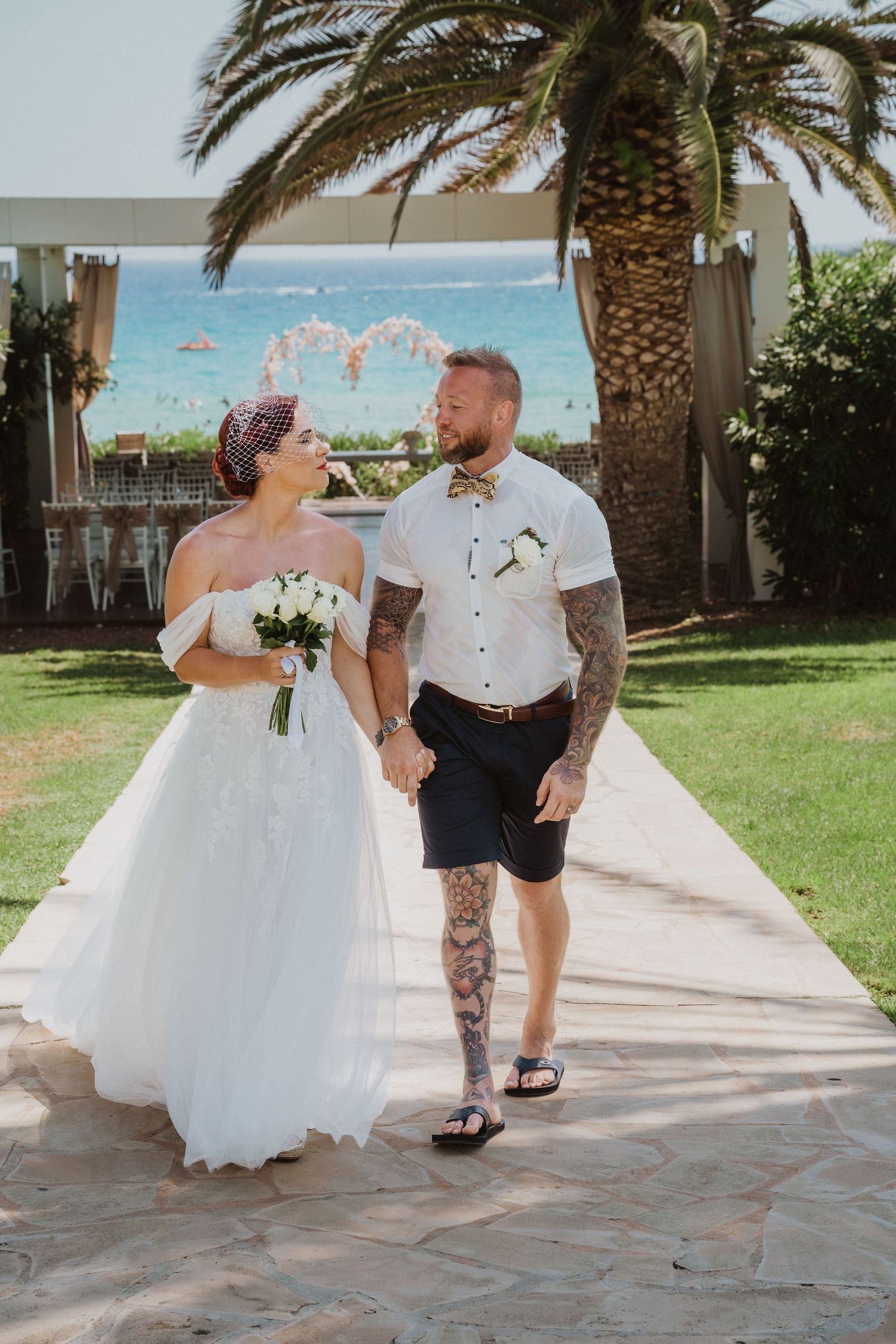 Newlyweds walking hand in hand on stone pathway with palm trees and ocean backdrop.