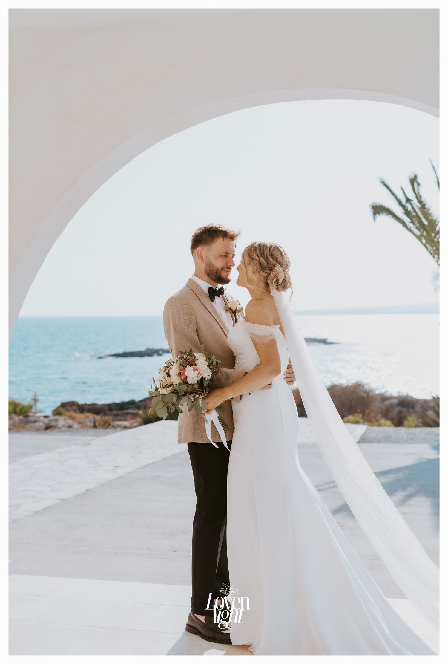 Romantic wedding portrait under white arch with Mediterranean Sea and palm tree in background.