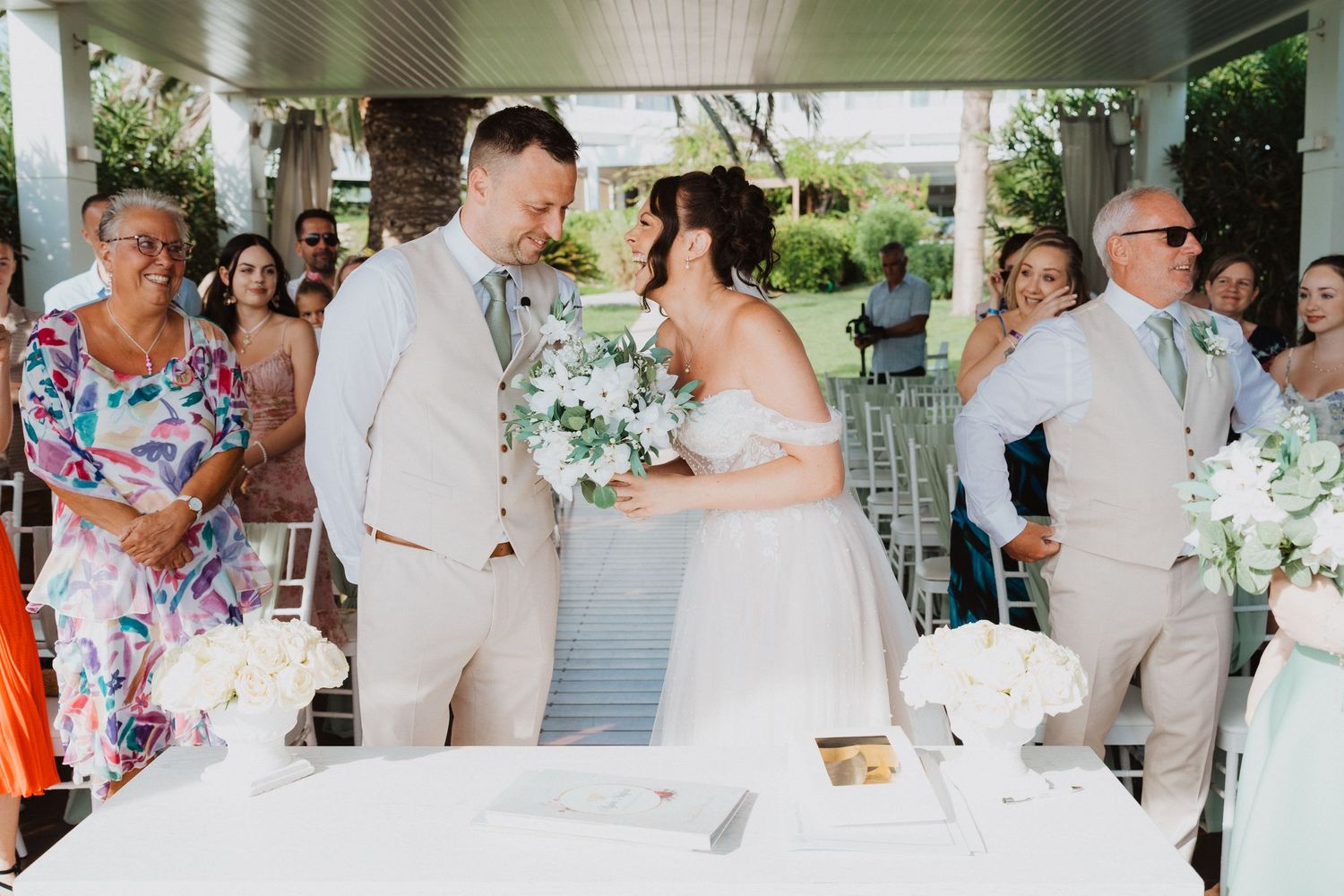 A wedding couple shares a joyful moment during their outdoor ceremony surrounded by guests and floral decorations.