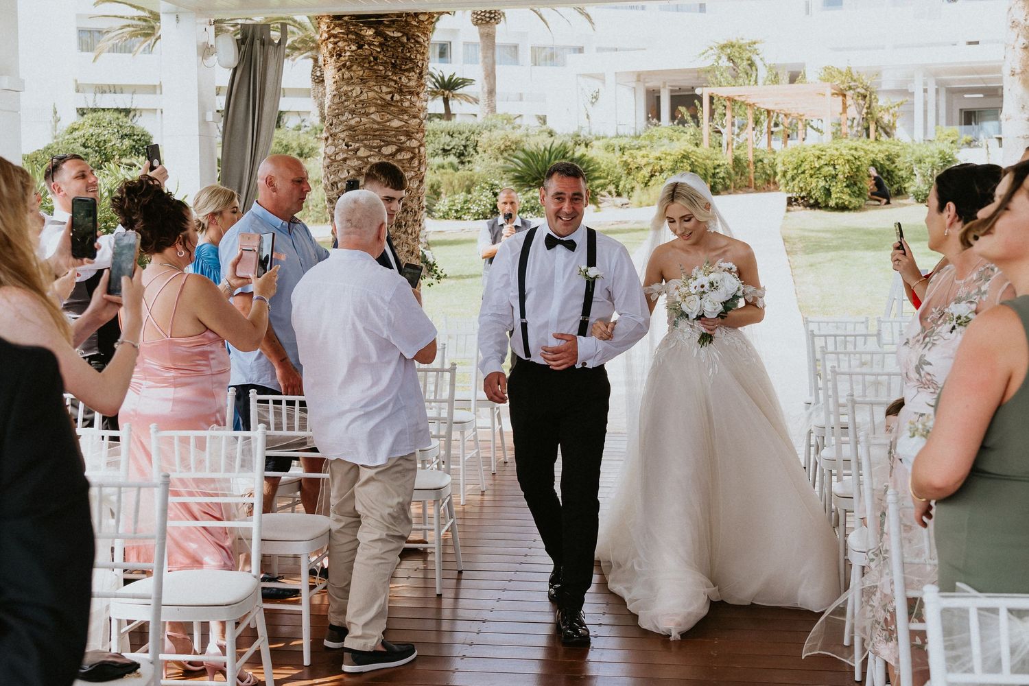 Wedding ceremony guests seated in white chairs on a covered outdoor patio.