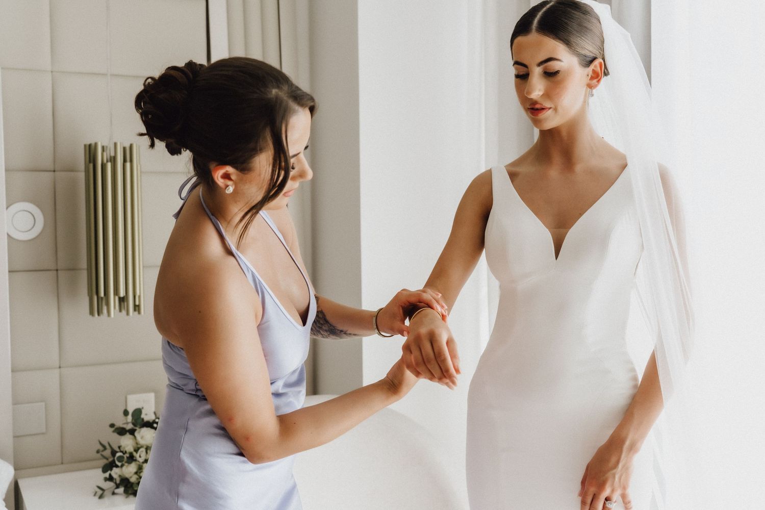 A person in lavender dress helps adjust the sleeve of a wedding gown.