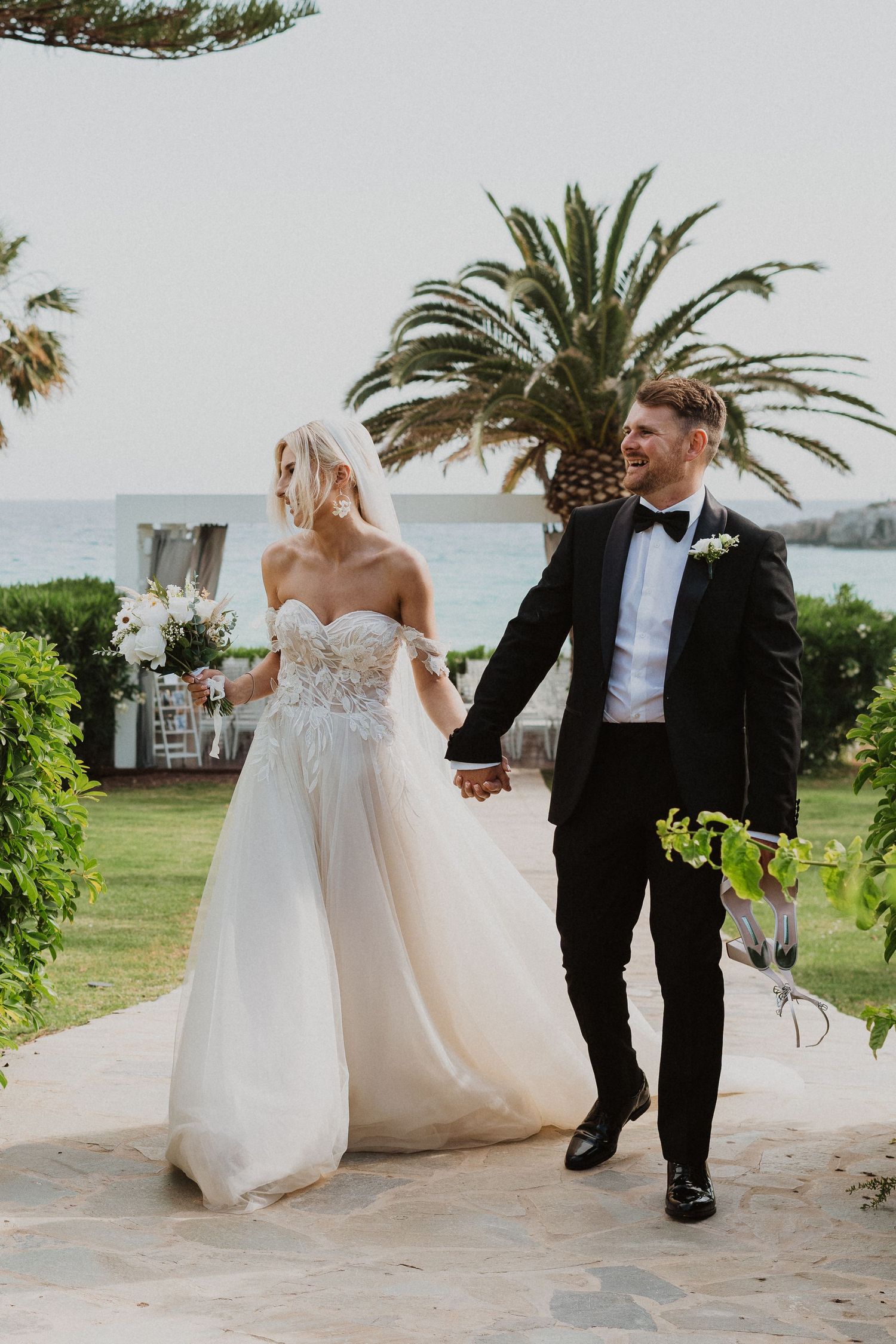 Newlyweds walk hand-in-hand through tropical garden setting at coastal wedding venue.
