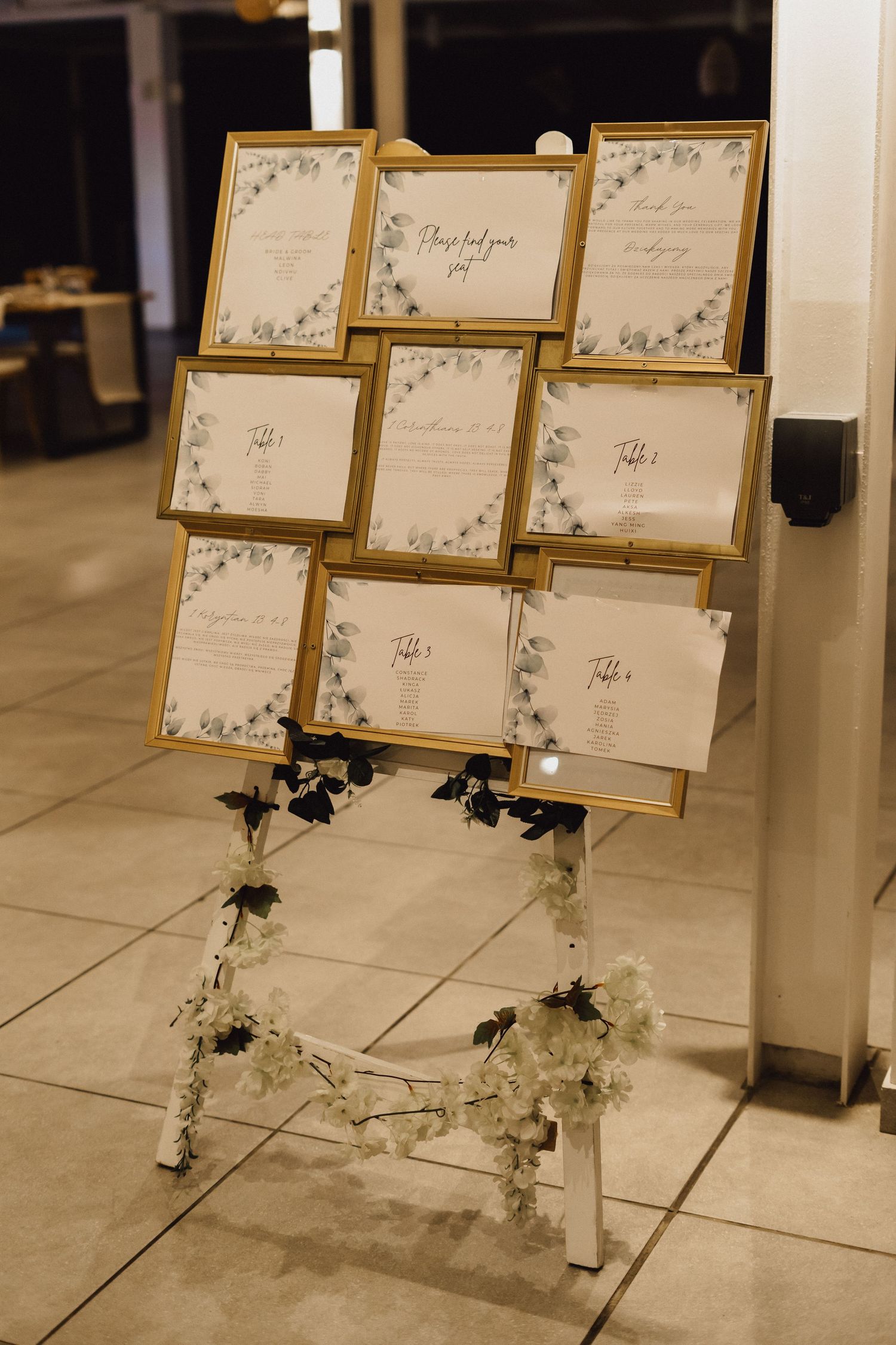 A wedding seating chart display with gold frames and floral decorations on a white stand.