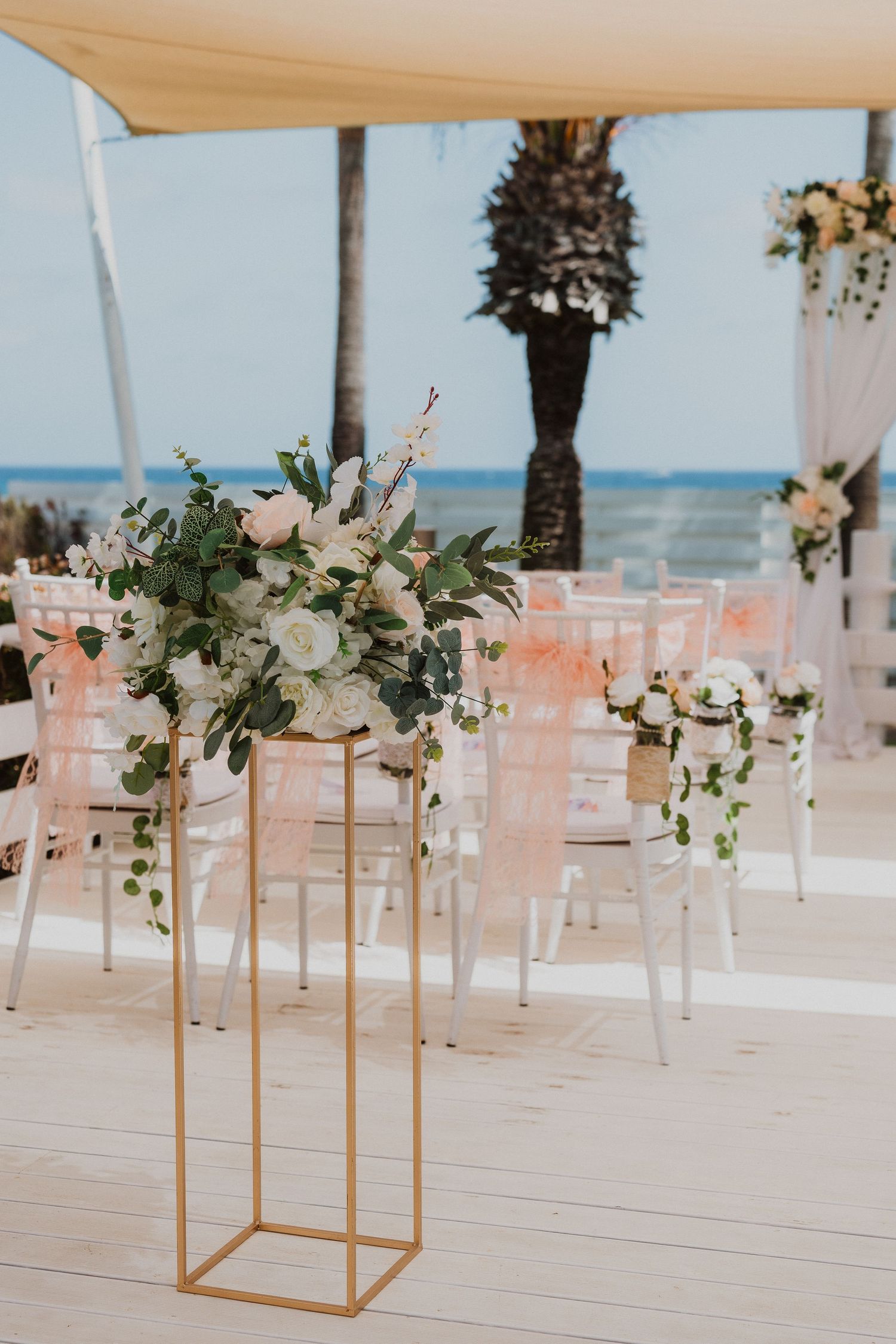 Elegant beach wedding setup with gold floral stands and ghost chairs under a white canopy by the ocean.