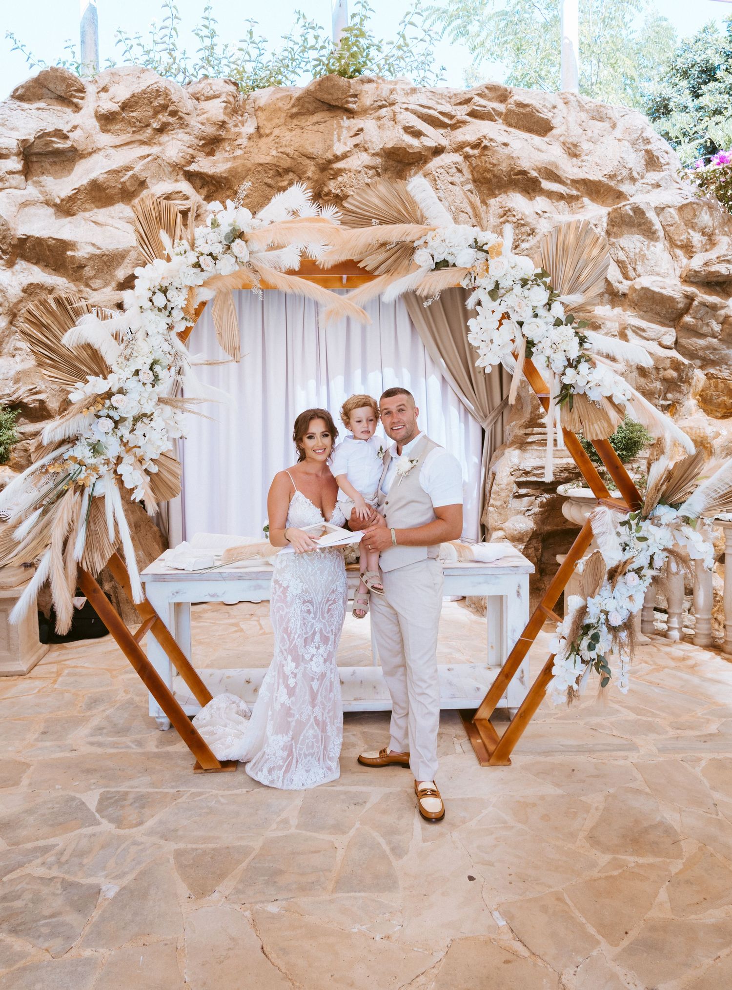 Wedding photo featuring a beautifully decorated arch with dried palms and white florals against a textured stone backdrop.