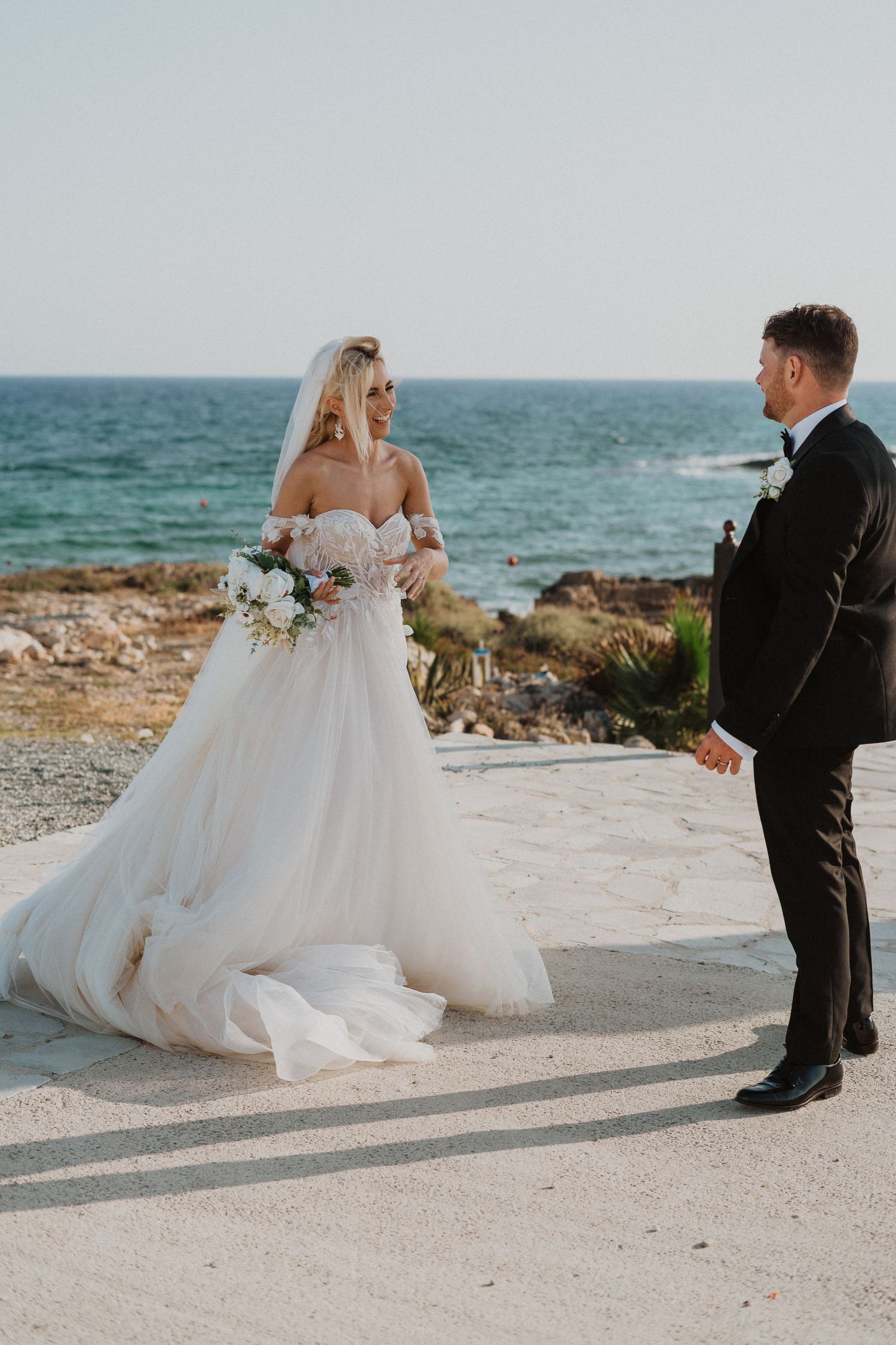Bride in strapless gown and groom share romantic moment by Mediterranean Sea.