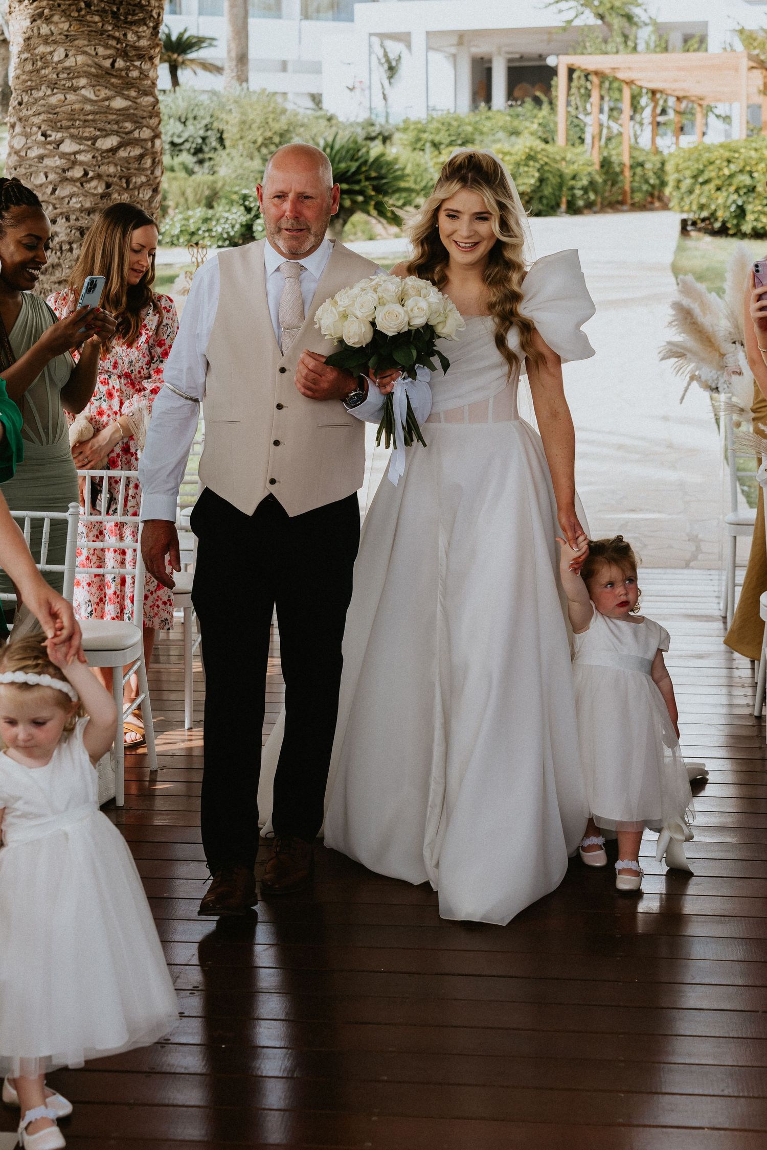 Wedding procession enters outdoor ceremony space with palm trees and white chairs.