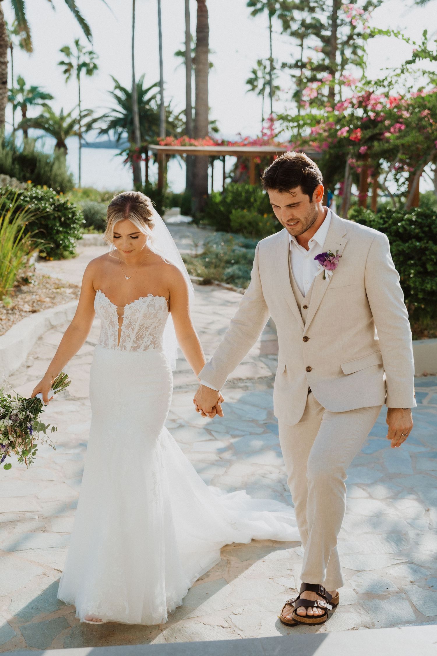 A joyful wedding couple walking hand-in-hand along a palm-lined tropical pathway.