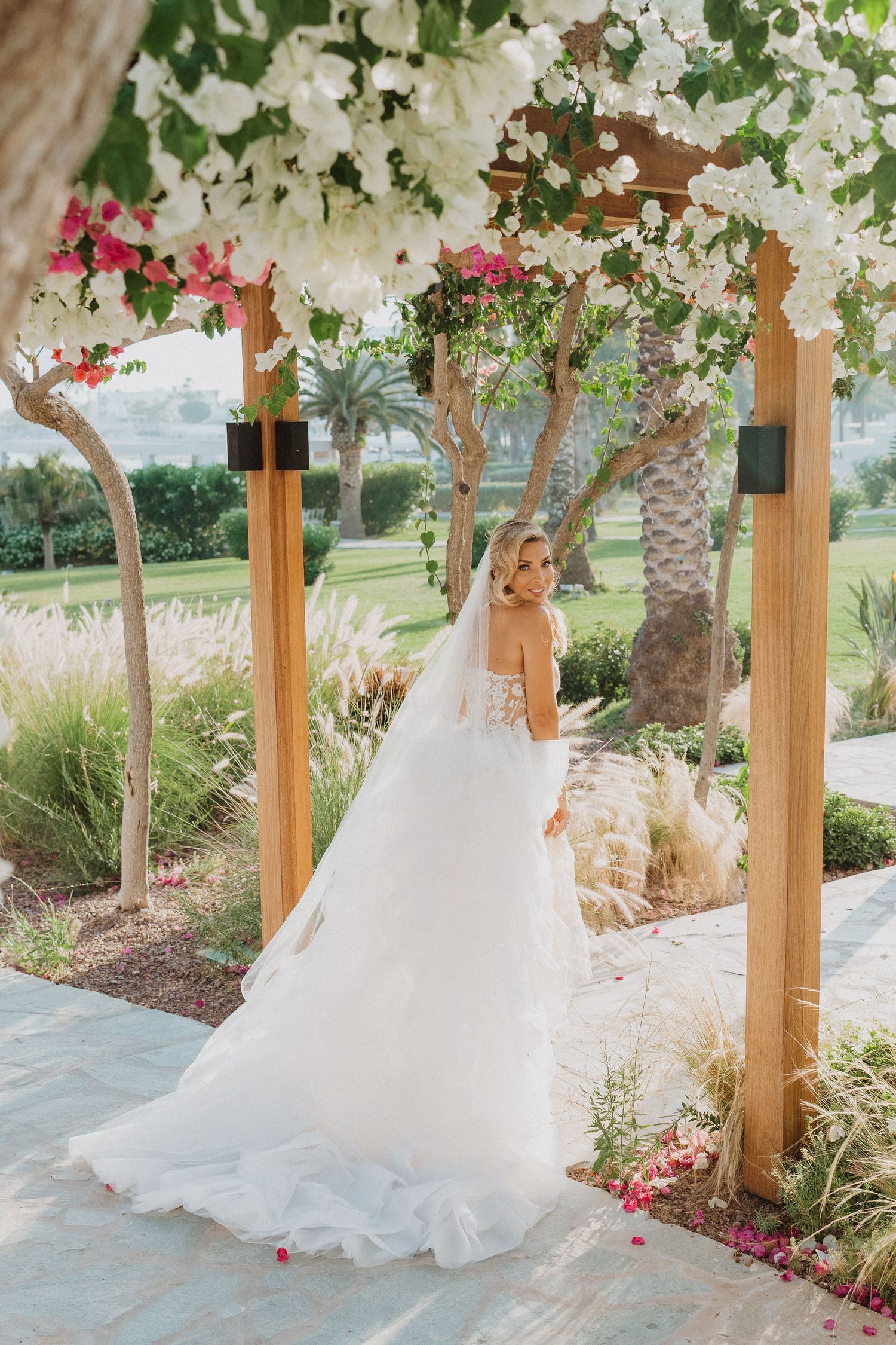 A white wedding dress flows elegantly under a wooden pergola adorned with cascading white flowers and pink blooms.