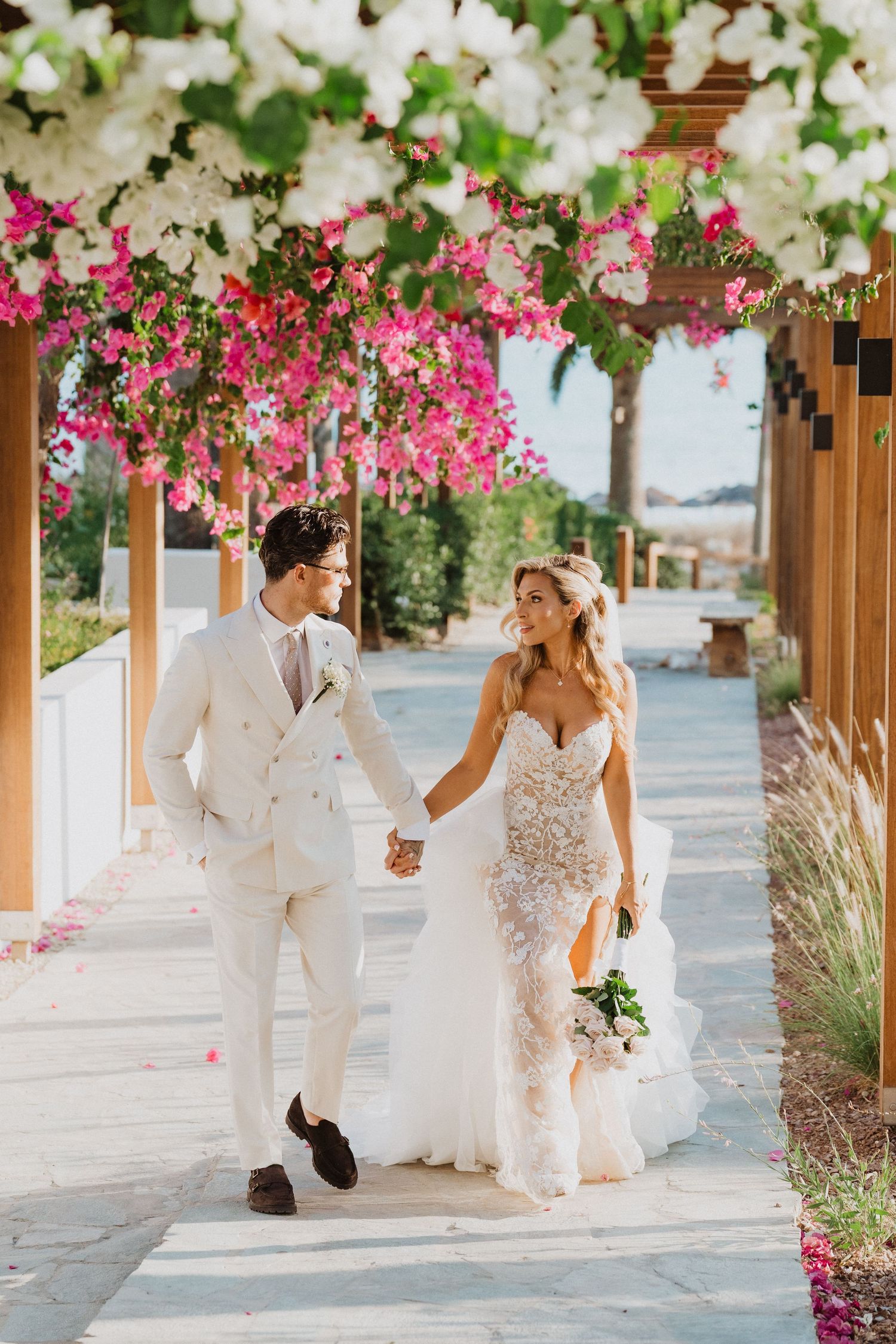 Bride and groom walk together under a covered walkway adorned with vibrant pink bougainvillea flowers and white blooms.