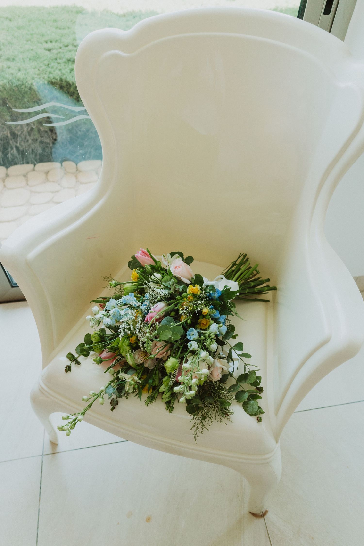 A delicate floral bouquet with pink and yellow blooms rests on an elegant white upholstered armchair by a window.