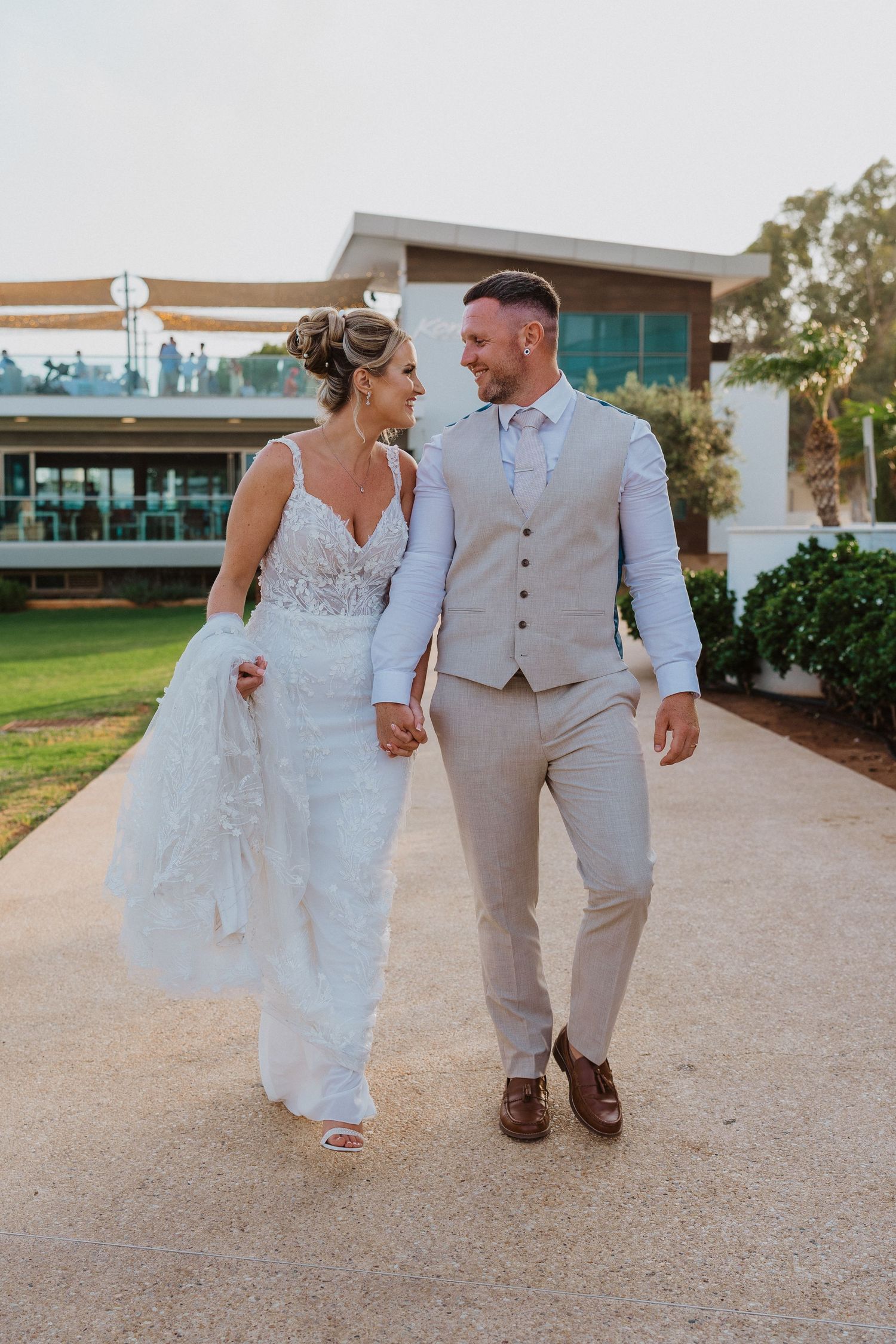 A couple in wedding attire walks together on a gravel path, with the bride in white and groom in tan suit.